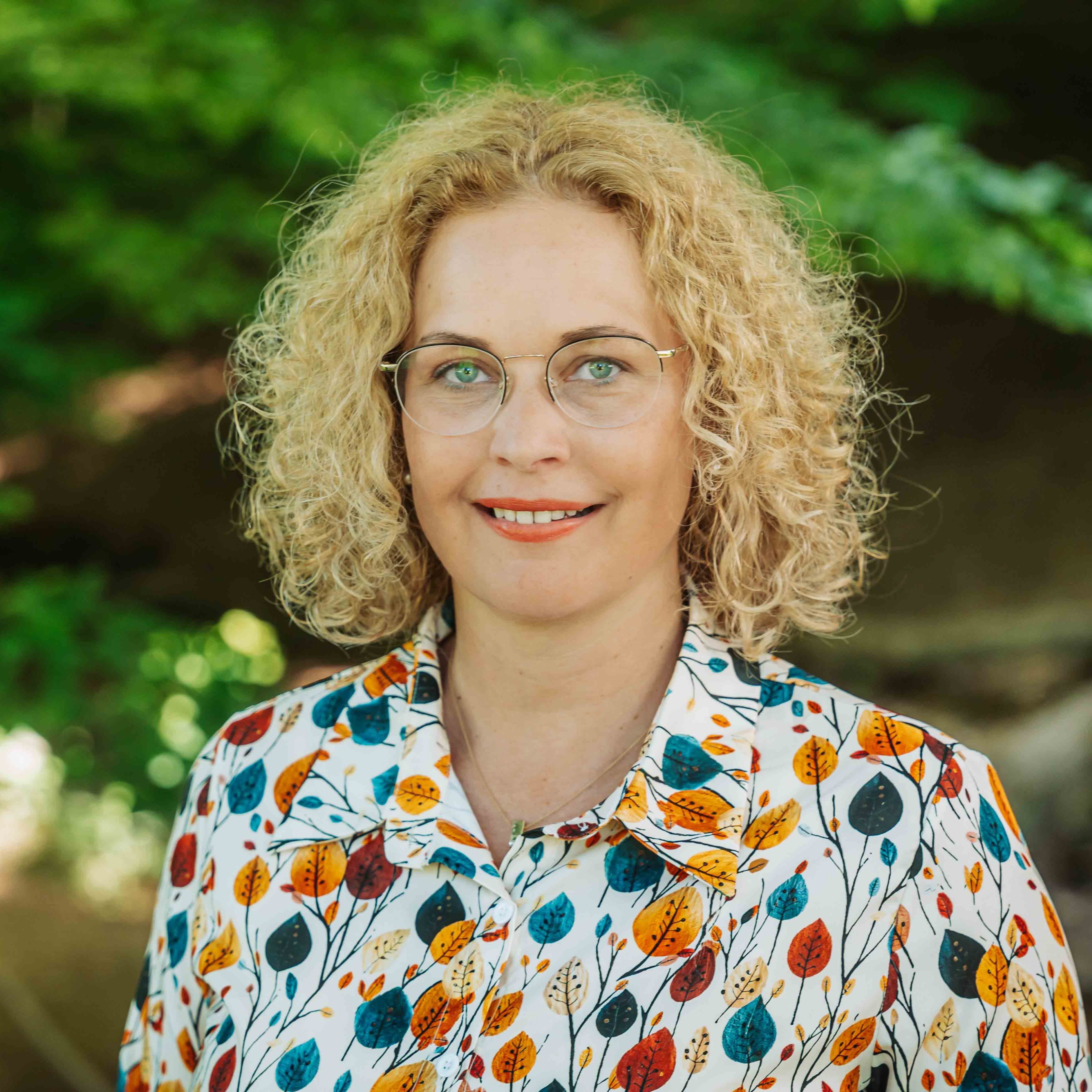 Alea Portrait einer Frau mit lockigem blondem Haar und bunter Bluse im Grünen