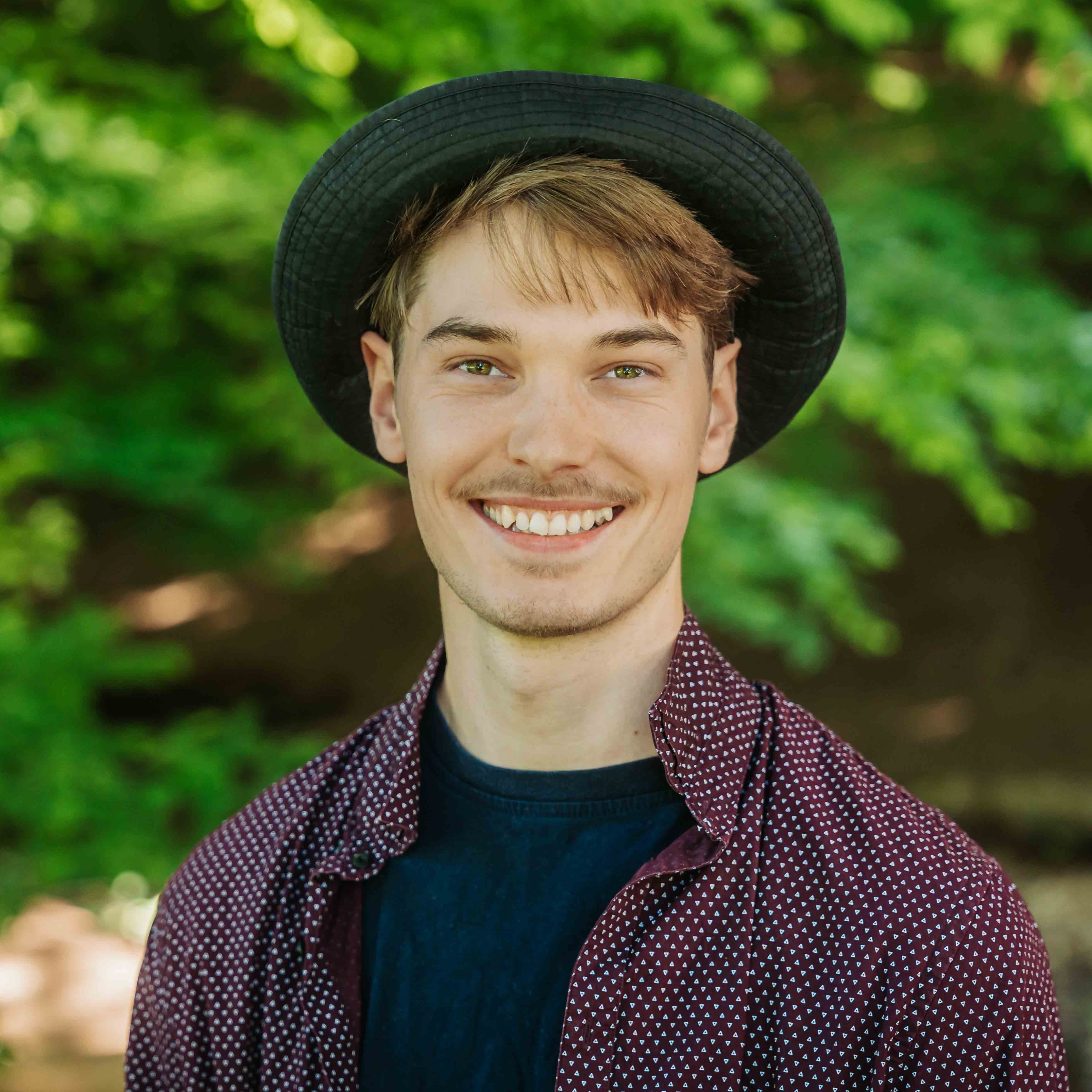 Alea Portrait eines jungen Mannes mit Hut und gemusterter Hemdjacke vor grünem Hintergrund