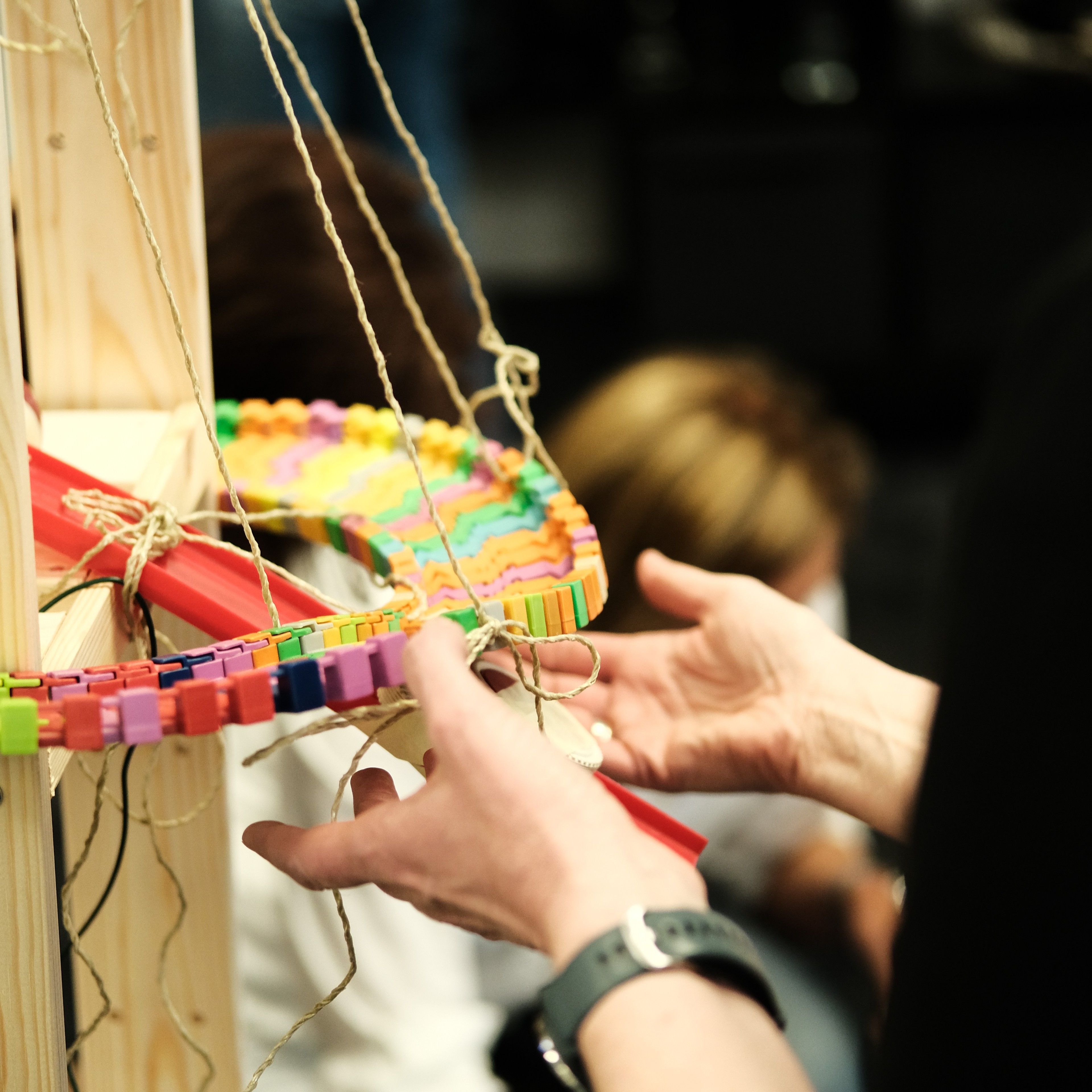 Person baut mit bunten Bausteinen und Schnüren an einer Holzkonstruktion, Detailaufnahme der Hände