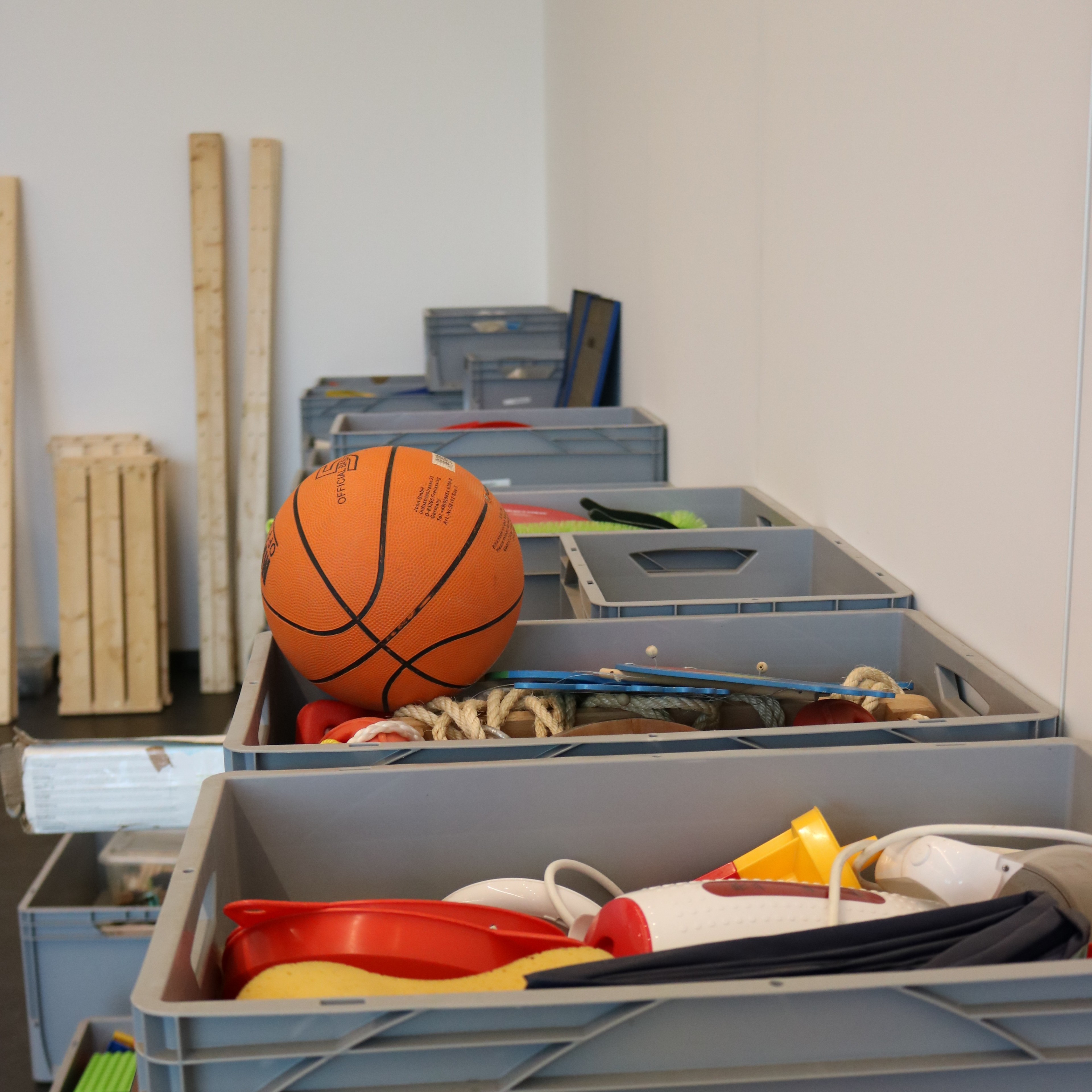 Mehrere graue Kisten mit Sportgeräten, darunter ein Basketball, in einem Sportraum