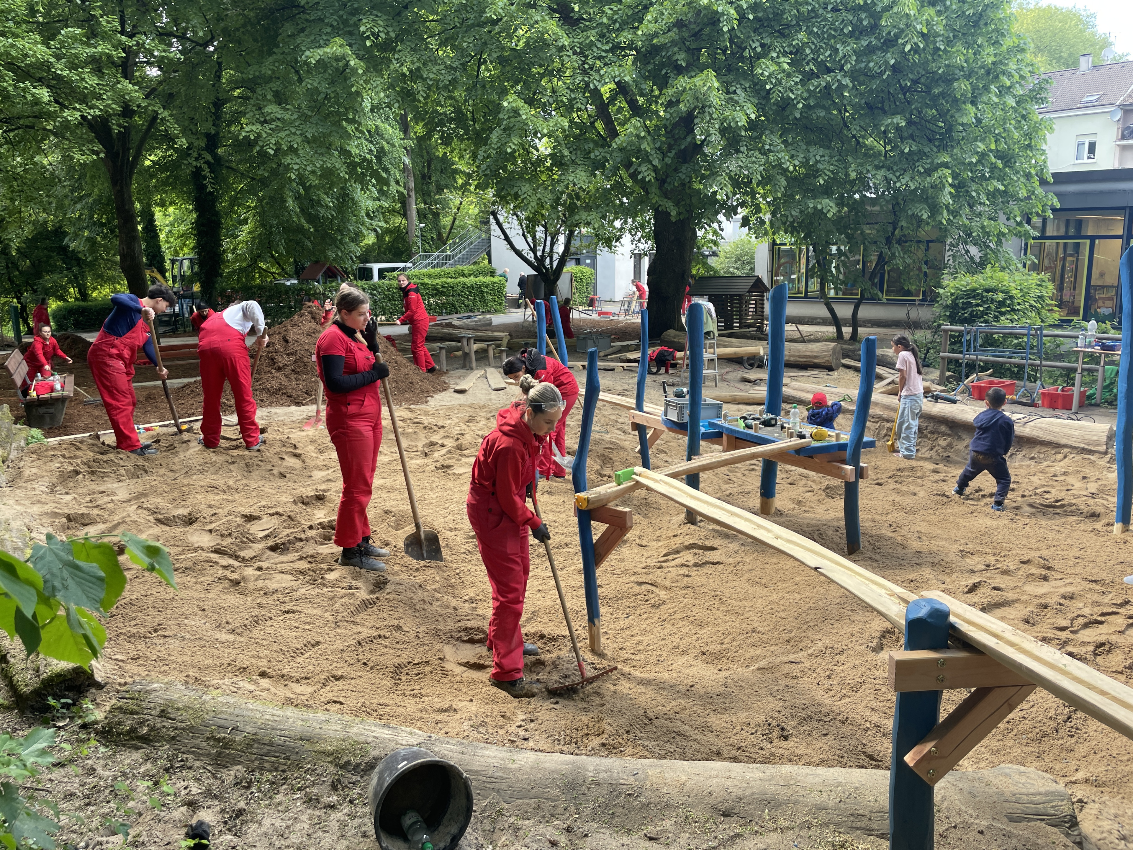 Personen in roter Arbeitskleidung gestalten einen Spielplatz im Sand mit Holz- und Kletterelementen