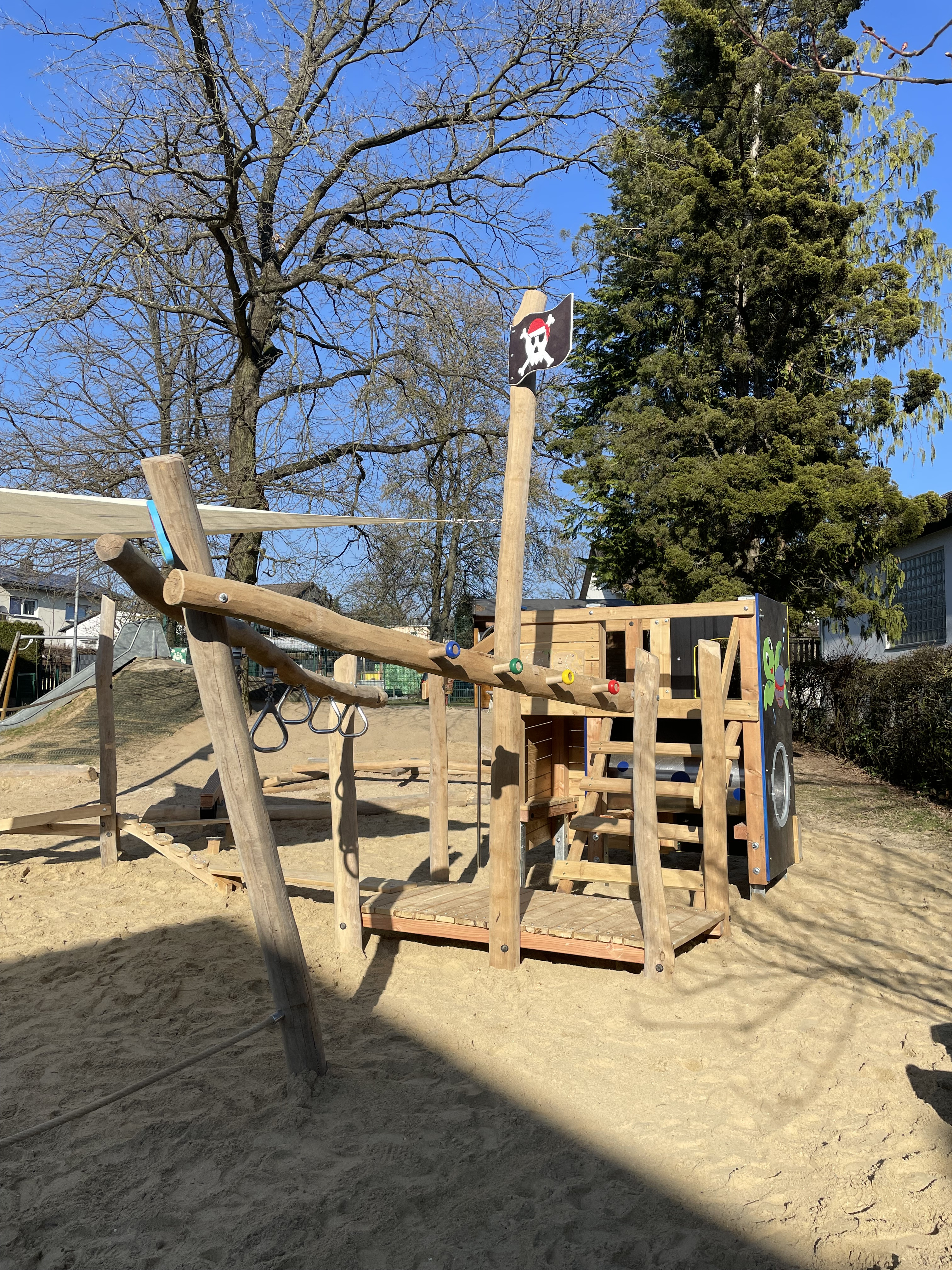 Holzspielplatz mit Klettergerüst und Piratenflagge im Sand unter blauem Himmel
