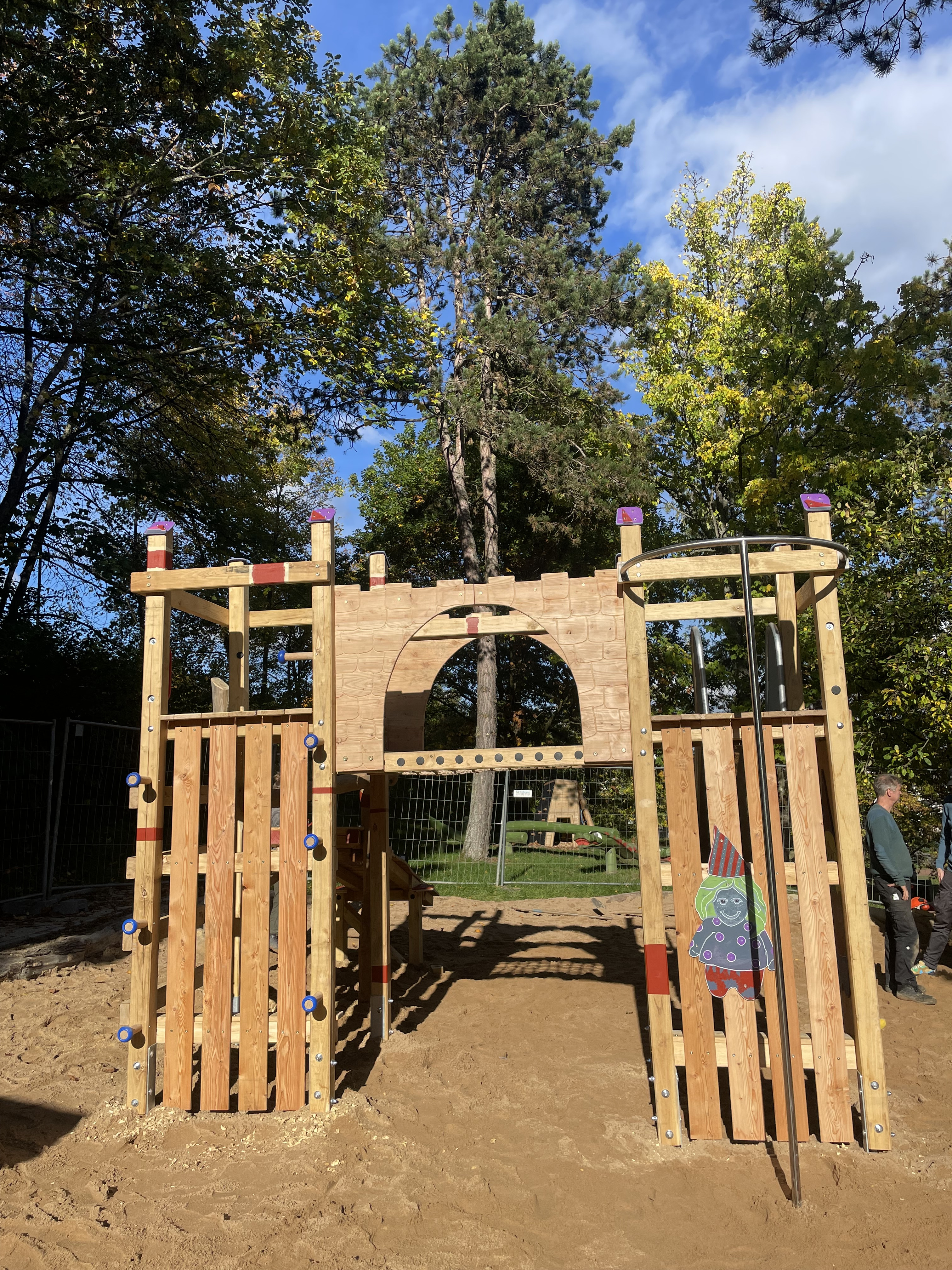 Holzspielplatz im Freien mit Kletterturm und Sandfläche bei sonnigem Wetter