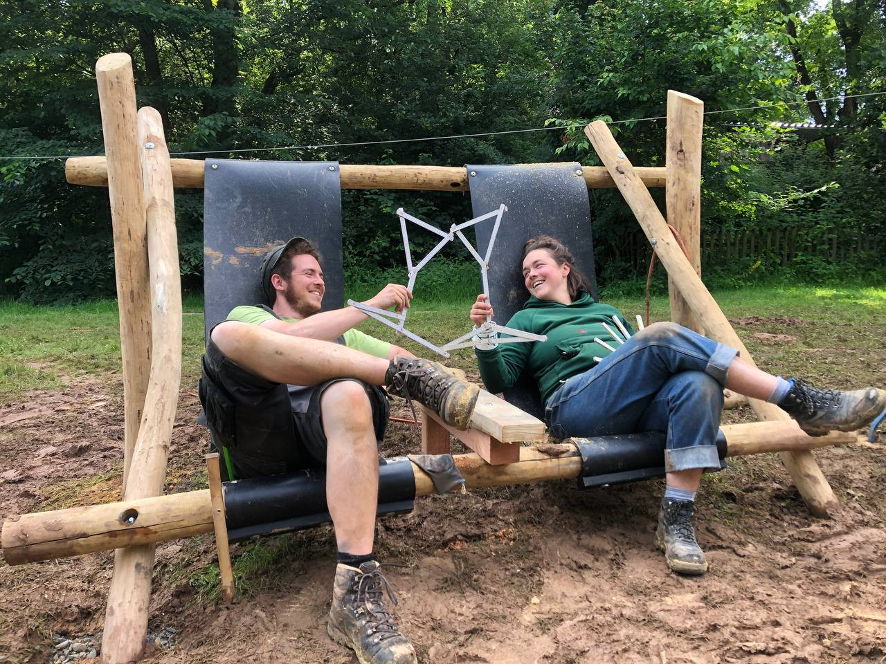 Zwei Personen entspannen auf einer selbstgebauten Holzschaukel im Freien und halten eine geometrische Konstruktion aus Stäben in den Händen.