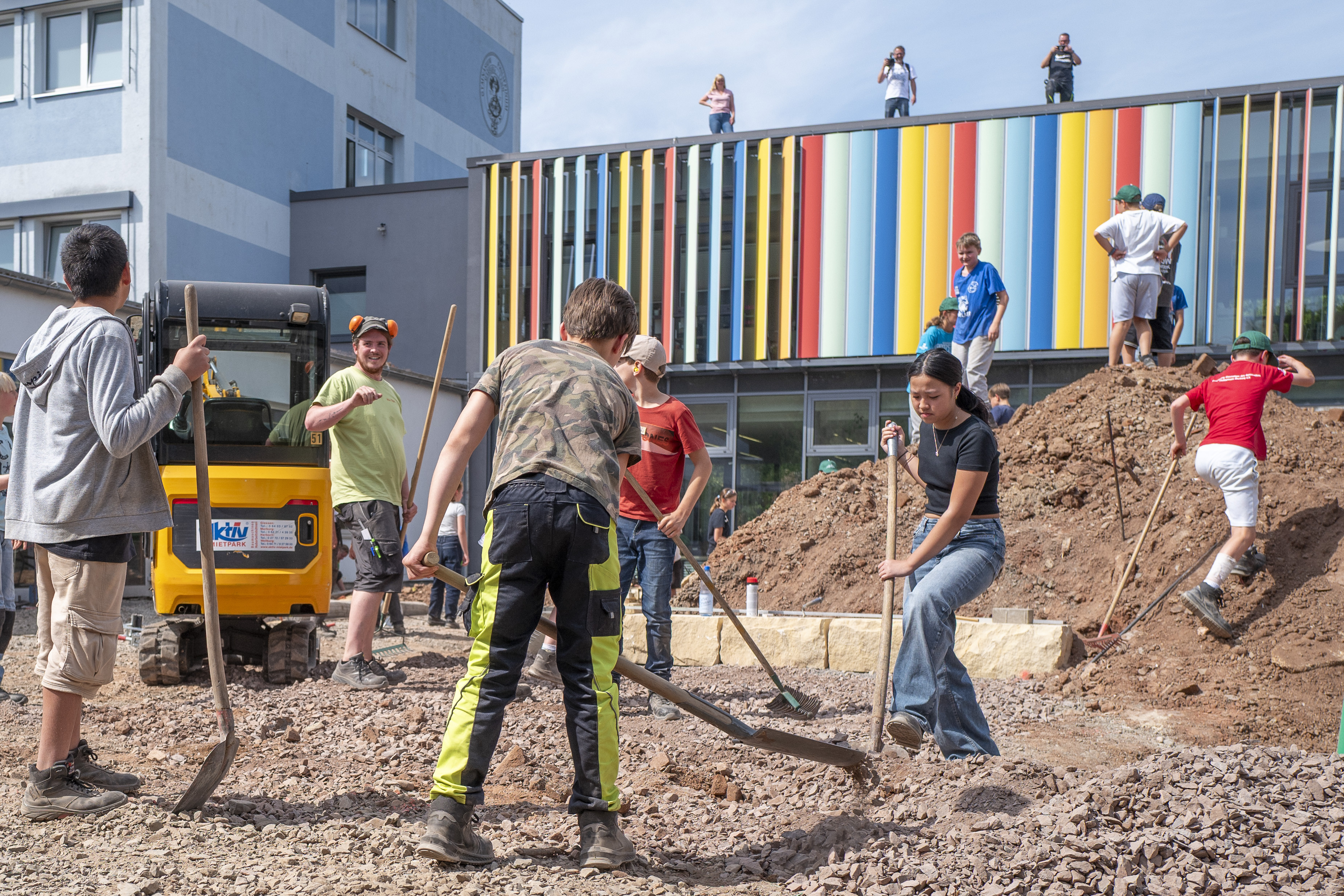 Schüler arbeiten gemeinsam auf einer Baustelle vor einem modernen, bunt gestalteten Schulgebäude