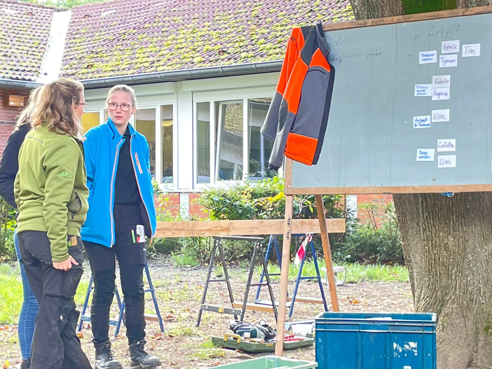 Zwei Personen bei einer Outdoor-Besprechung vor einer Tafel mit Werkzeug und Arbeitsmaterialien im Freien