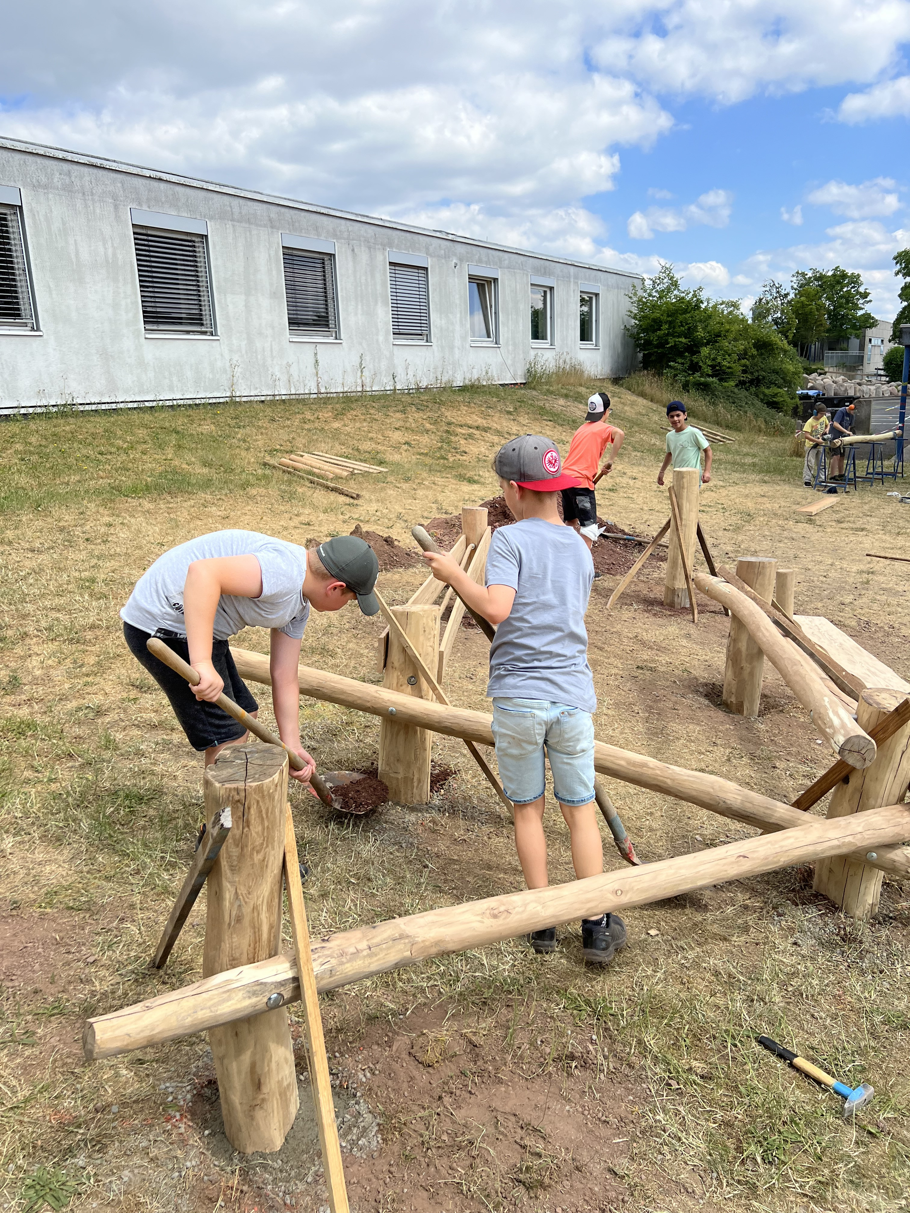 Kinder bauen gemeinsam einen Spielplatz aus Holz auf einer Wiese vor einem Schulgebäude