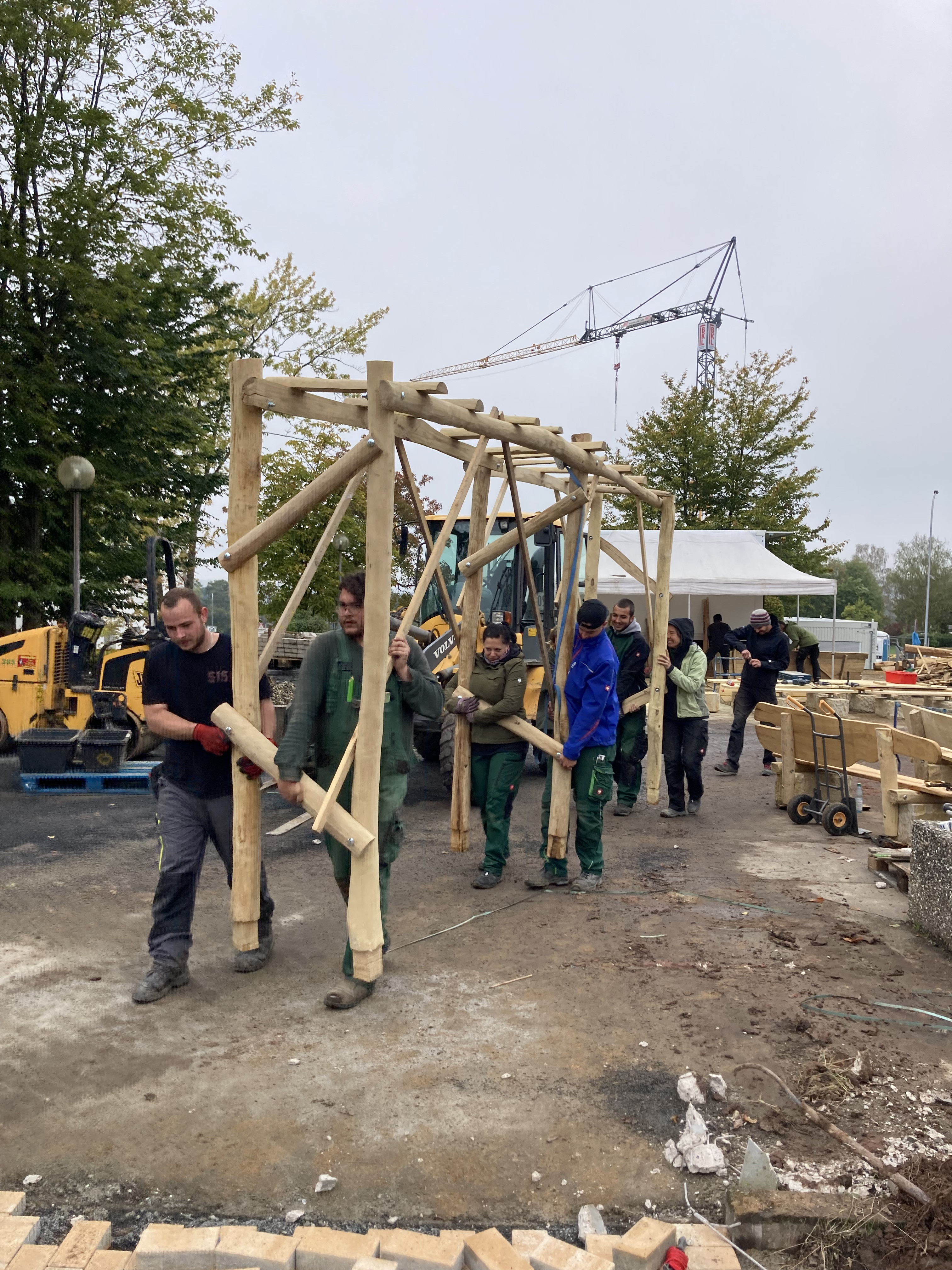 Mehrere Personen tragen gemeinsam eine große Holzkonstruktion auf einer Baustelle im Freien