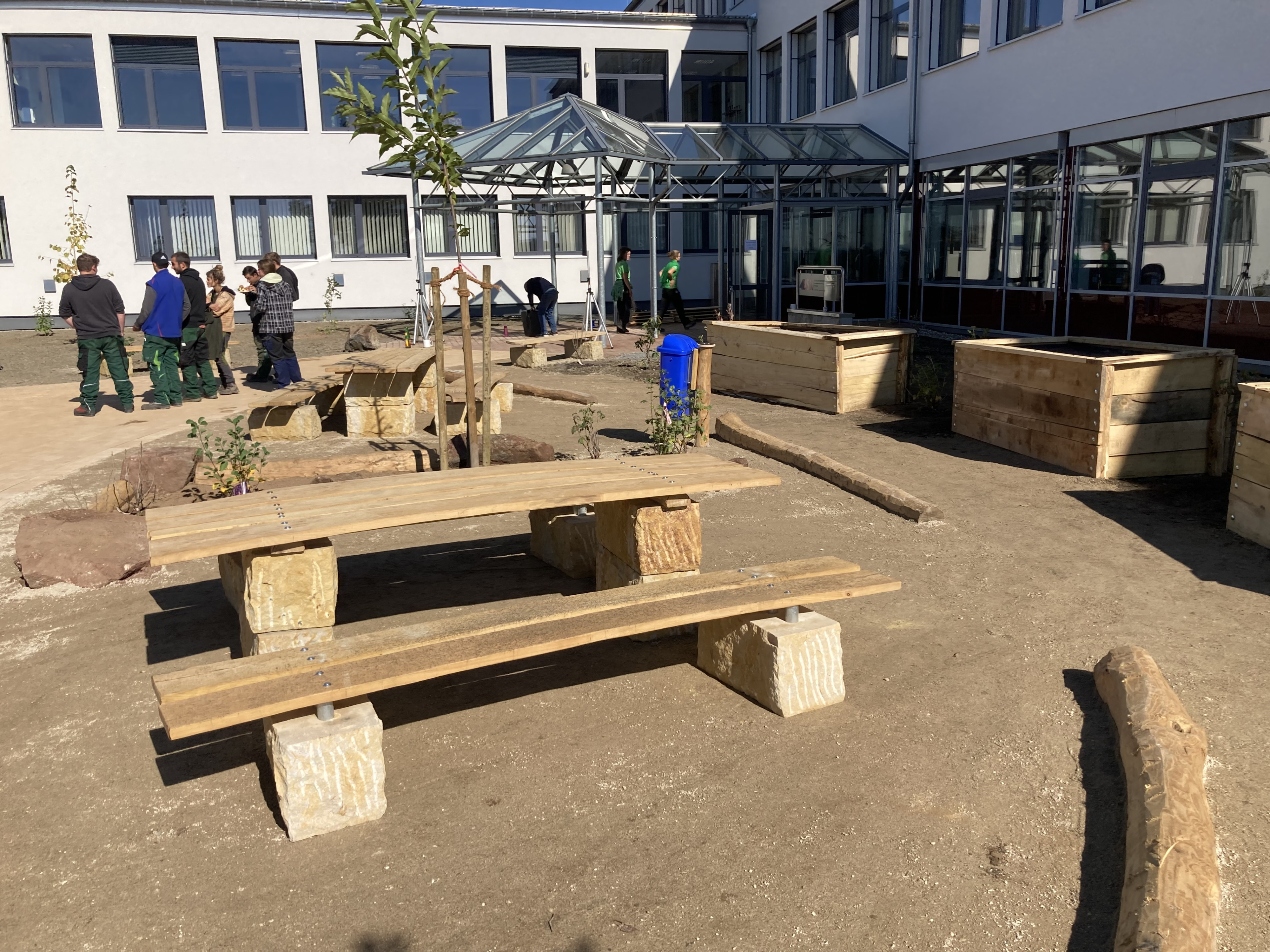 Innenhof mit neuen Holzbänken und Hochbeeten vor einem modernen Schulgebäude, mehrere Personen im Hintergrund