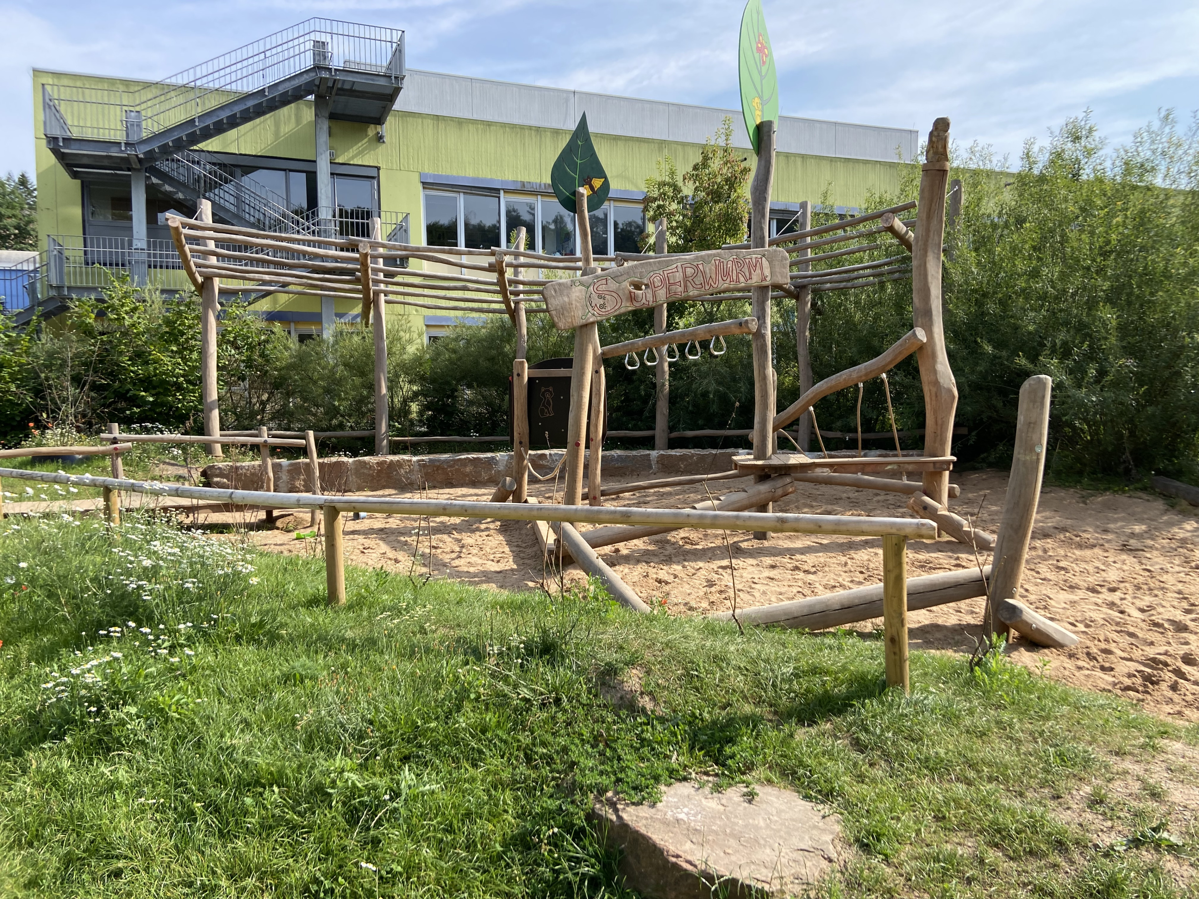Moderner Spielplatz aus Holz mit Sandfläche und Klettergerüst namens Superwurm vor einem Schulgebäude