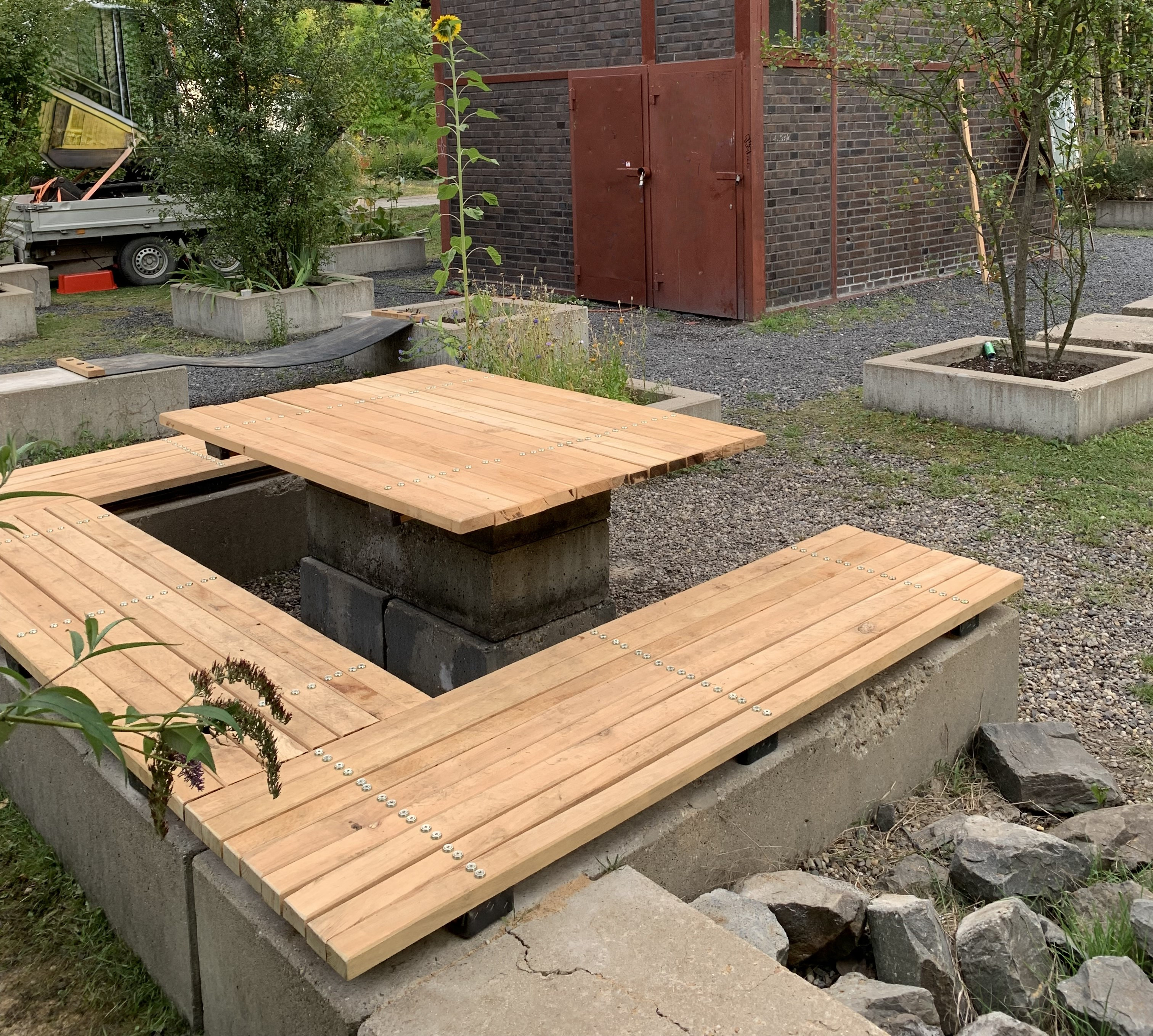 Moderner Sitzbereich aus Holz und Beton im urbanen Garten mit Sonnenblume und Backsteingebäude im Hintergrund