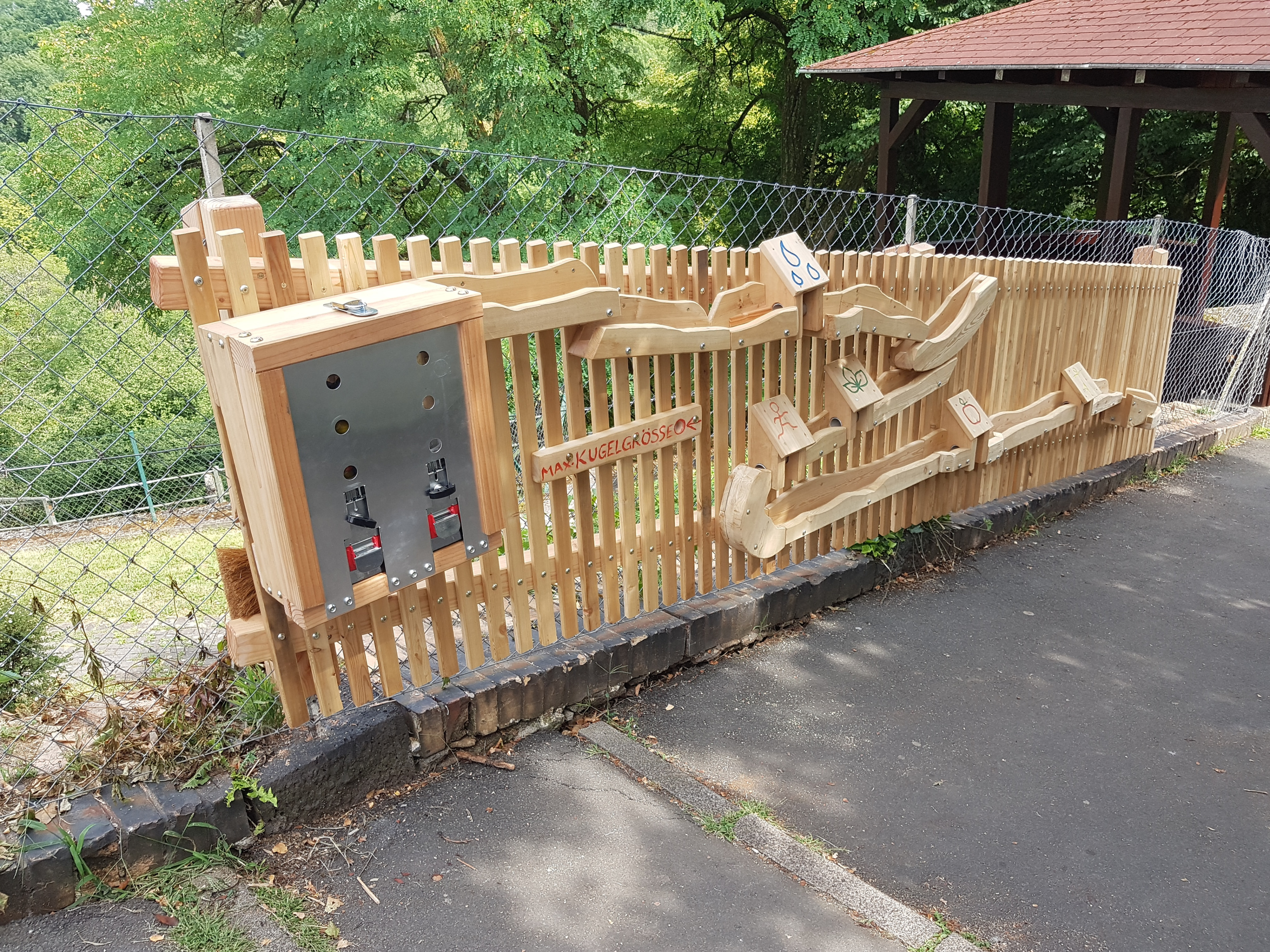 Holz-Murmelbahn im Freien an einem Zaun, mit verschiedenen Bahnelementen und Kugelspender