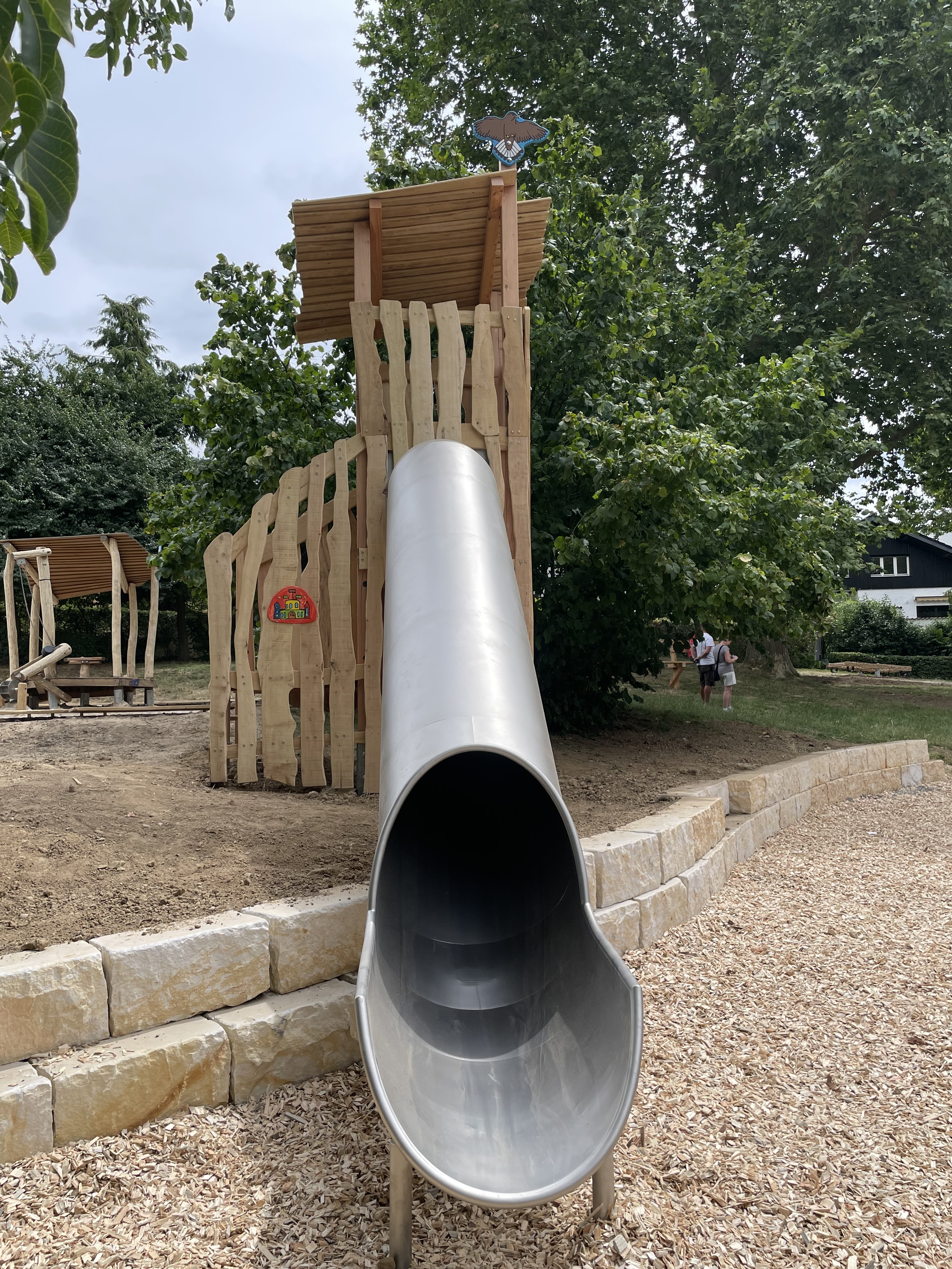 Große Edelstahlrutsche auf einem modernen Spielplatz mit Holzspielgeräten und Bäumen im Hintergrund