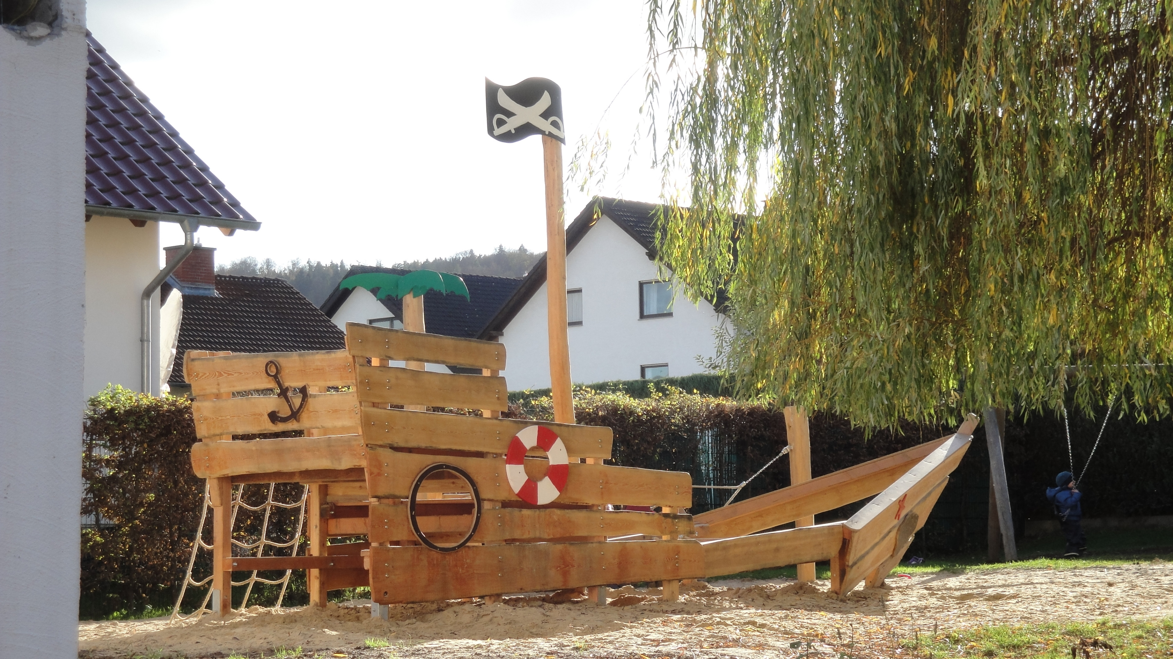 Holz-Piratenschiff auf einem Spielplatz mit Rettungsring und Anker, im Hintergrund Wohnhäuser und Bäume