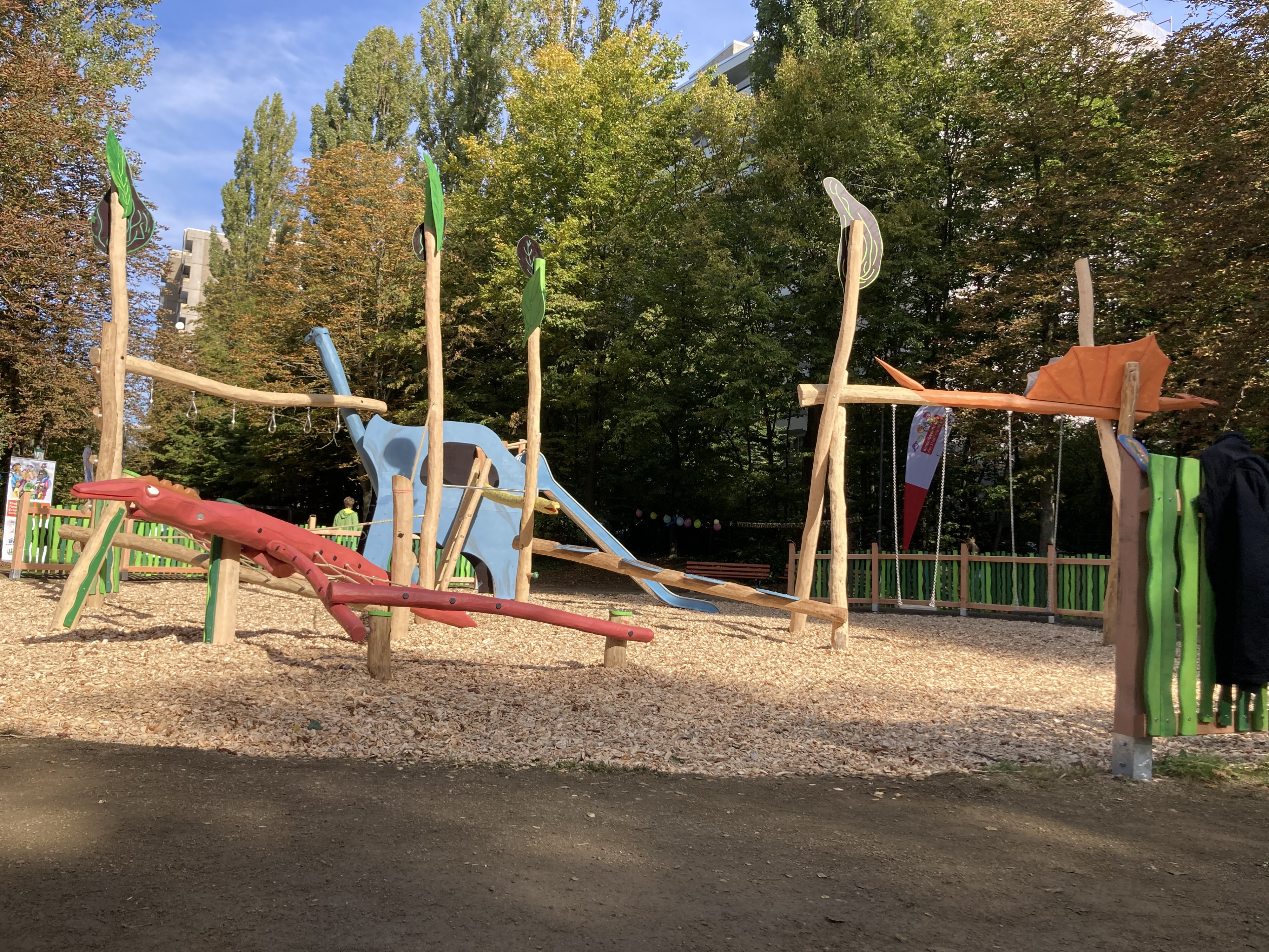Moderner Spielplatz mit Holzspielgeräten und Dinosaurier-Rutsche im Freien, umgeben von Bäumen