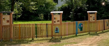 Holzzaun mit Infotafel und bemalten Elementen in einer grünen Parkanlage