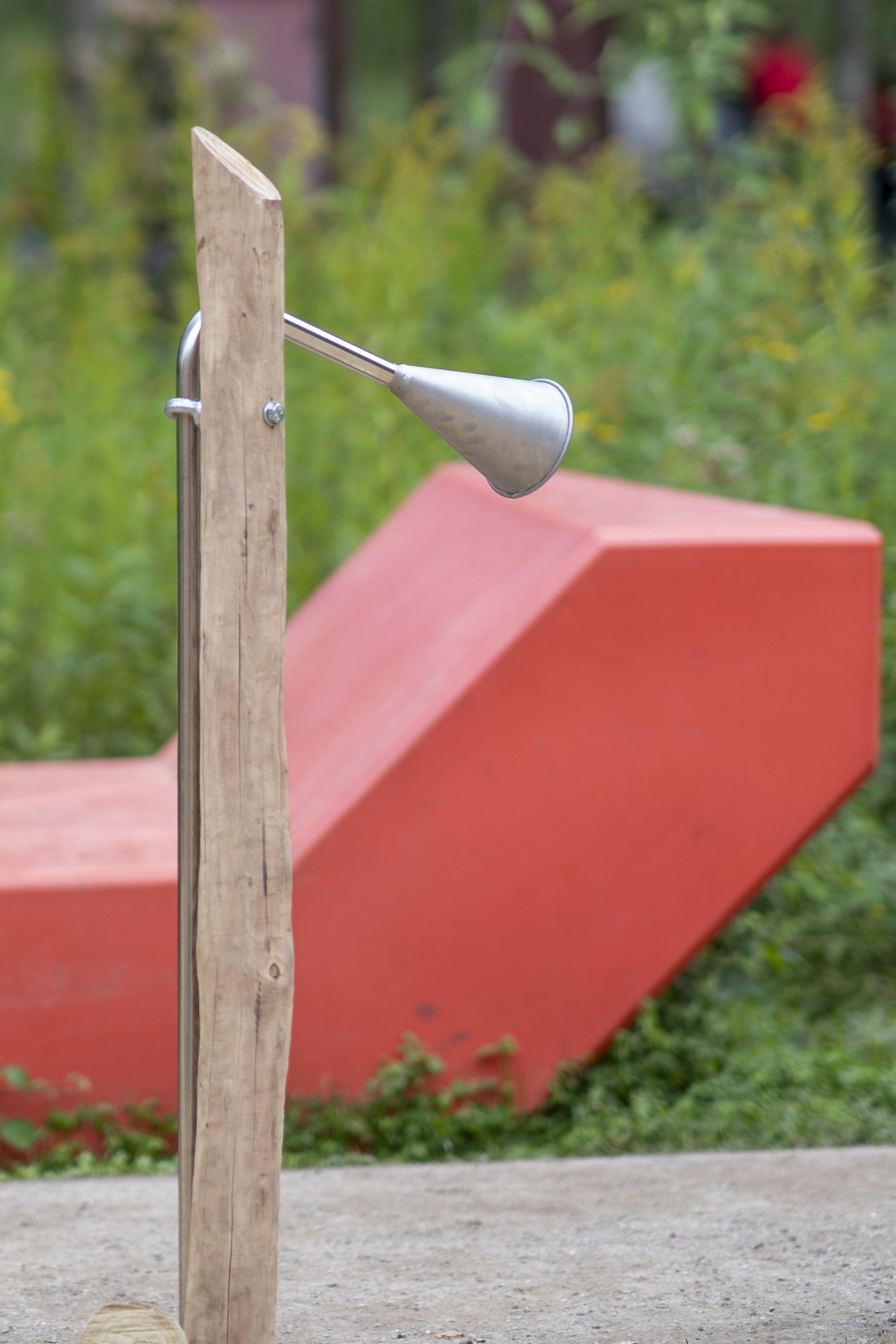Outdoor-Dusche aus Holz mit Metalduschkopf vor roter Sitzbank im Grünen