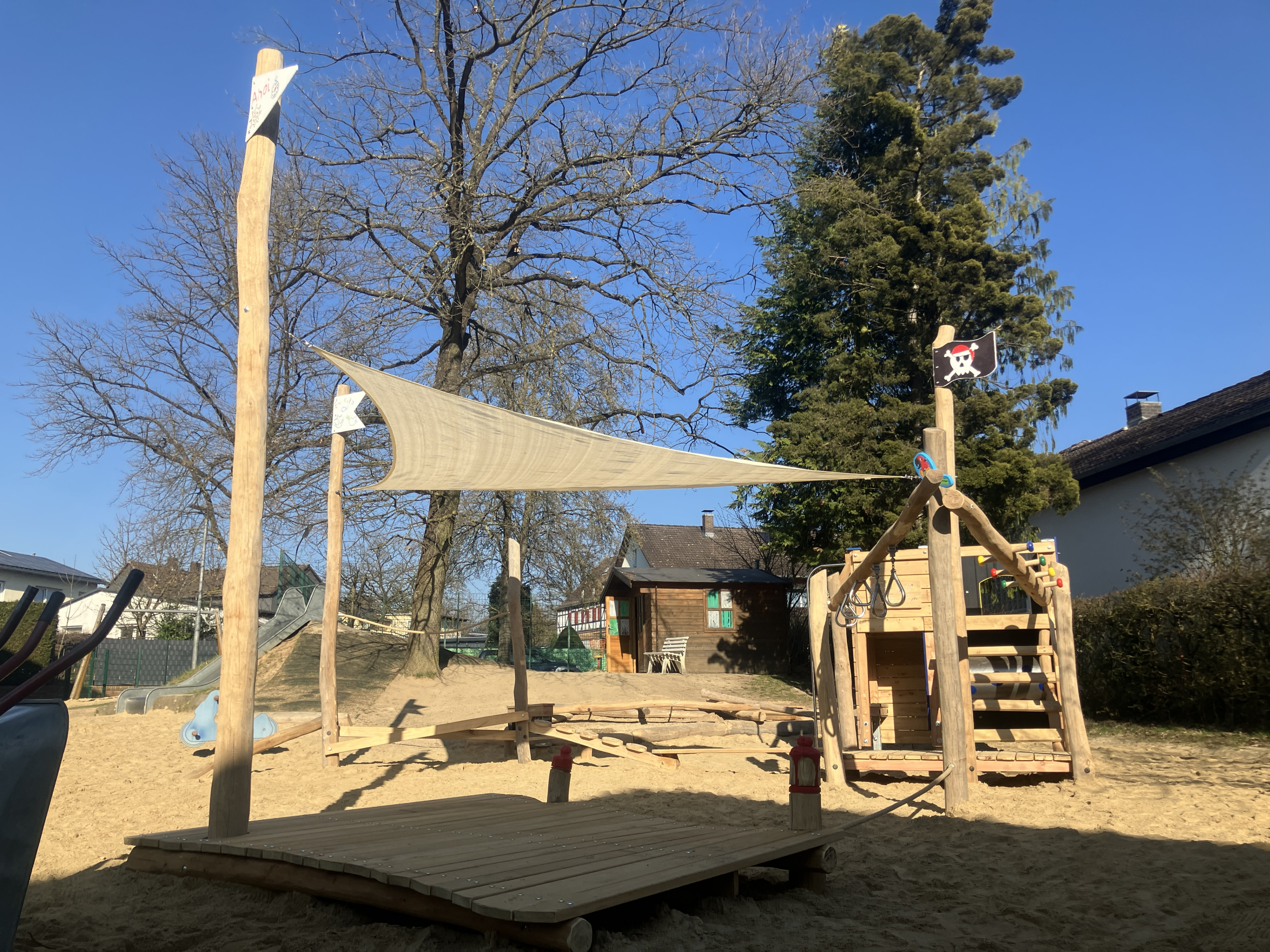 Moderner Spielplatz mit Holz-Piratenschiff, Sonnensegel und Sandfläche bei sonnigem Wetter