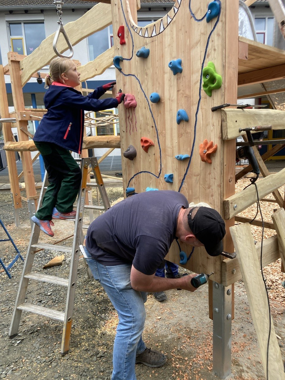 Erwachsener baut eine Kletterwand fr Kinder auf, whrend ein Kind auf einer Leiter steht und hilft