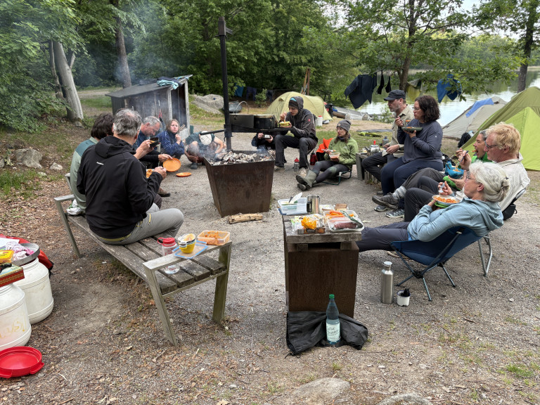 Gruppe von Menschen beim gemeinsamen Essen am Lagerfeuer auf einem Campingplatz im Grünen