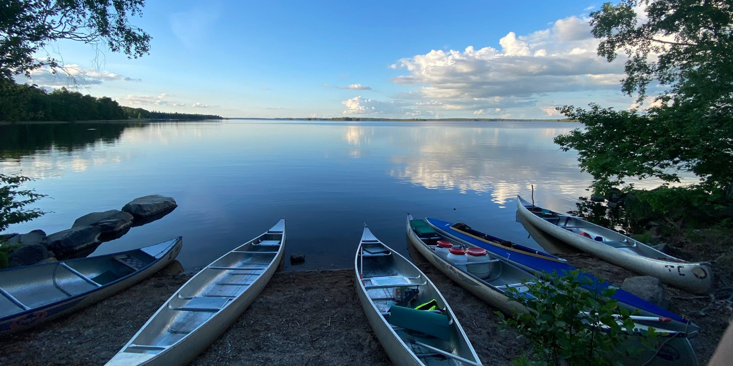 Mehrere Kanus am Ufer eines ruhigen Sees unter blauem Himmel mit Wolken