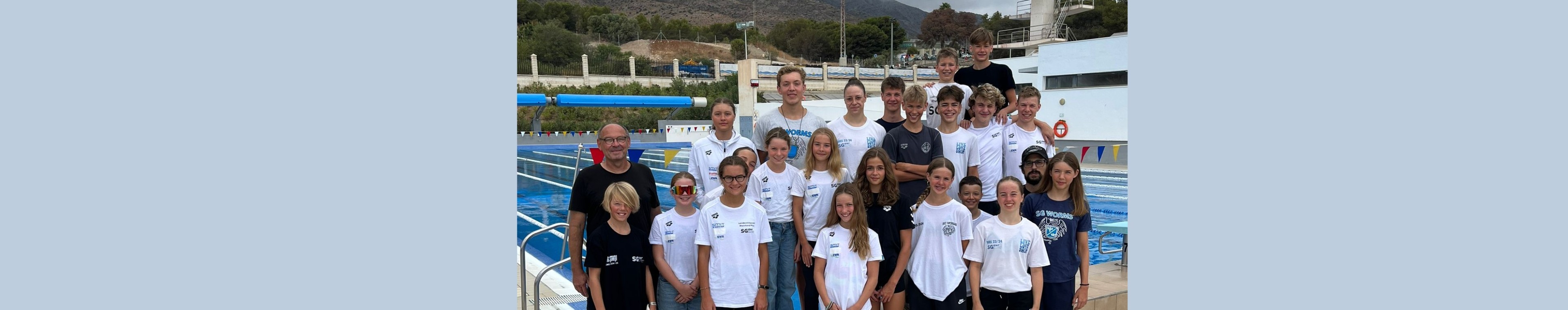 Gruppenfoto einer Schwimmgruppe am Beckenrand, aufgenommen im Freien mit Bergen im Hintergrund.