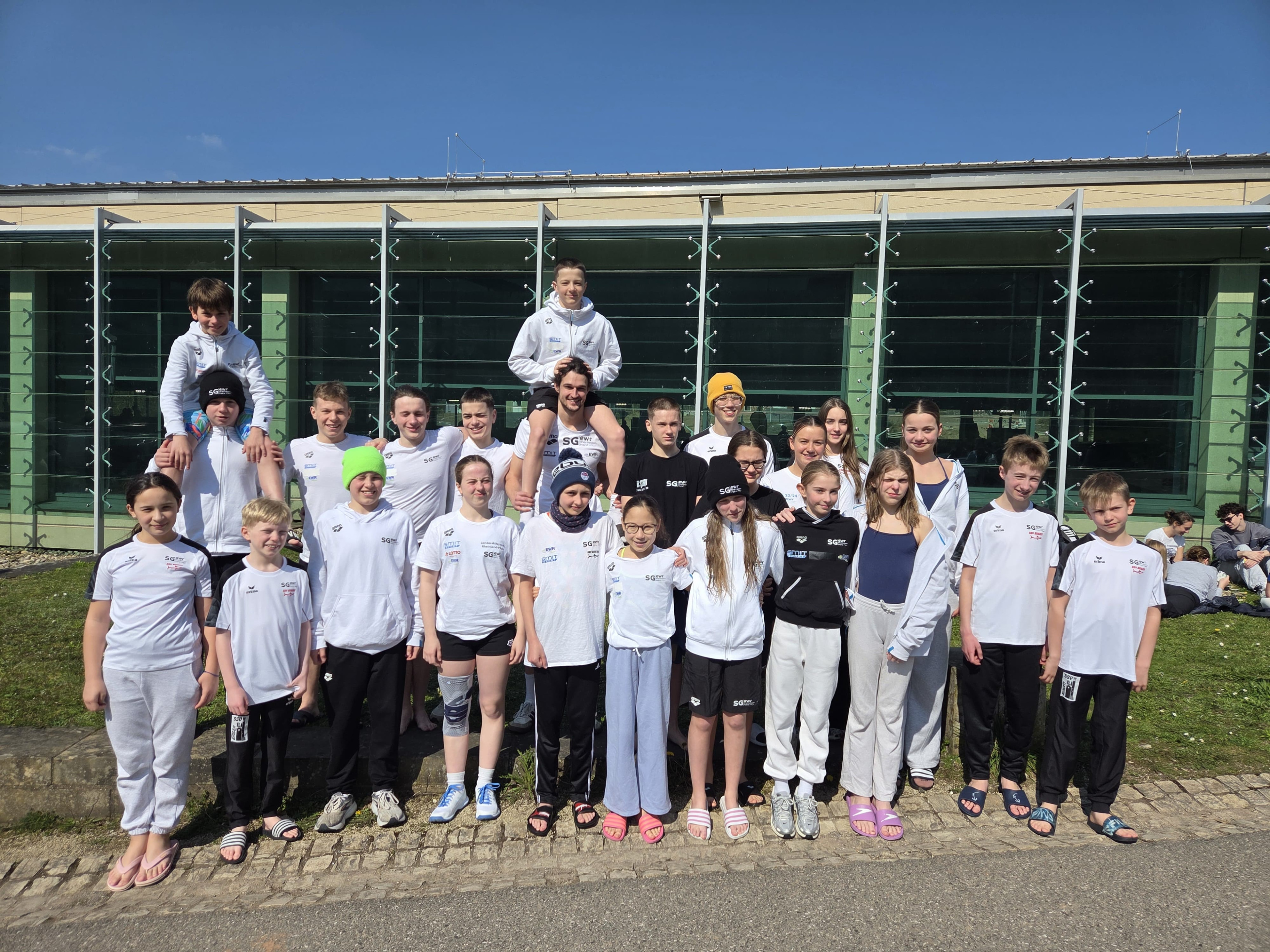 Gruppenfoto einer Jugend-Schwimmmannschaft vor einem modernen Gebäude bei sonnigem Wetter