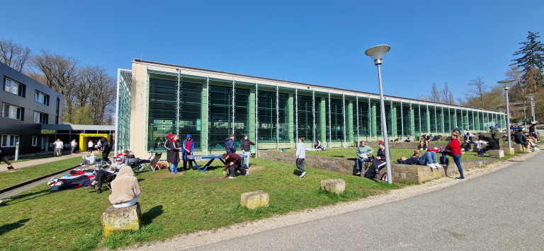 Menschen auf einer Grünfläche vor modernem Gebäude mit Glasfassade am 22.03.2026 Mehrere Personen verbringen Zeit auf einer Grünfläche vor einem modernen Gebäude mit großen Glasfassaden an einem sonnigen Tag.