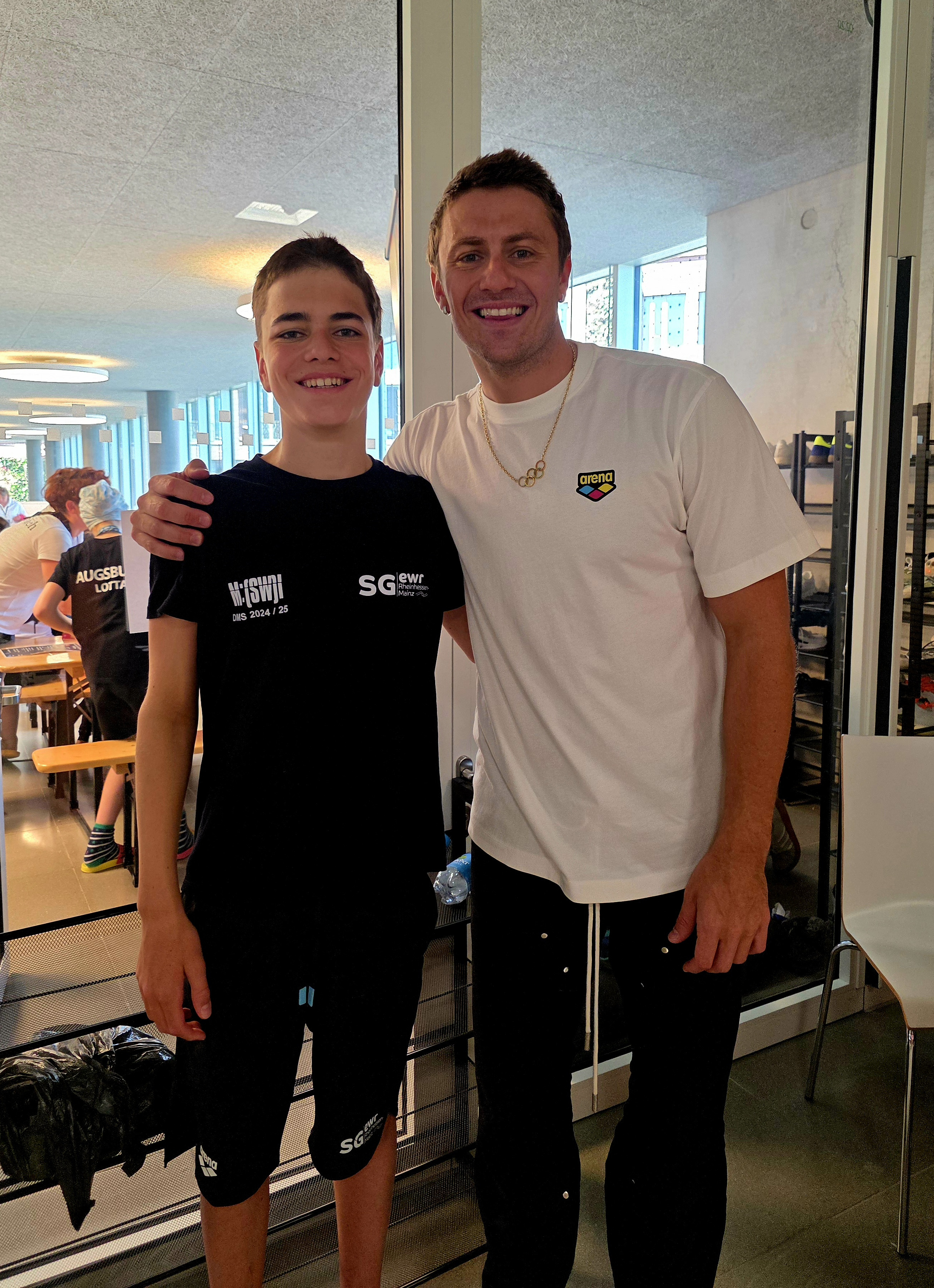 Zwei Personen bei einer Veranstaltung der SG EWR Mainz, einer trägt ein Arena T-Shirt, im Hintergrund weitere Teilnehmer