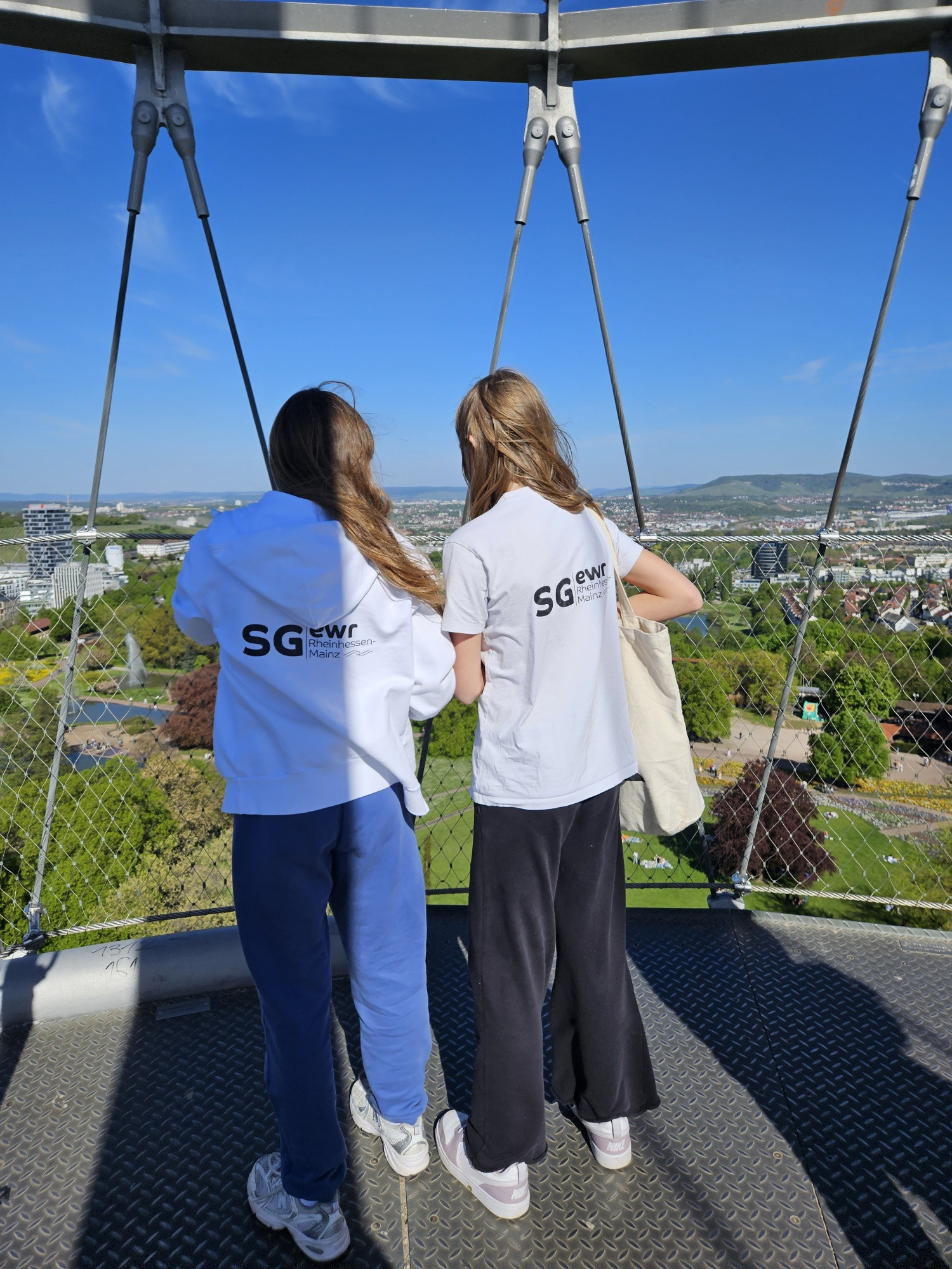 Zwei Personen mit SG EWR Rheinhessen-Mainz Kleidung genießen die Aussicht von einer Plattform über einer Stadtlandschaft