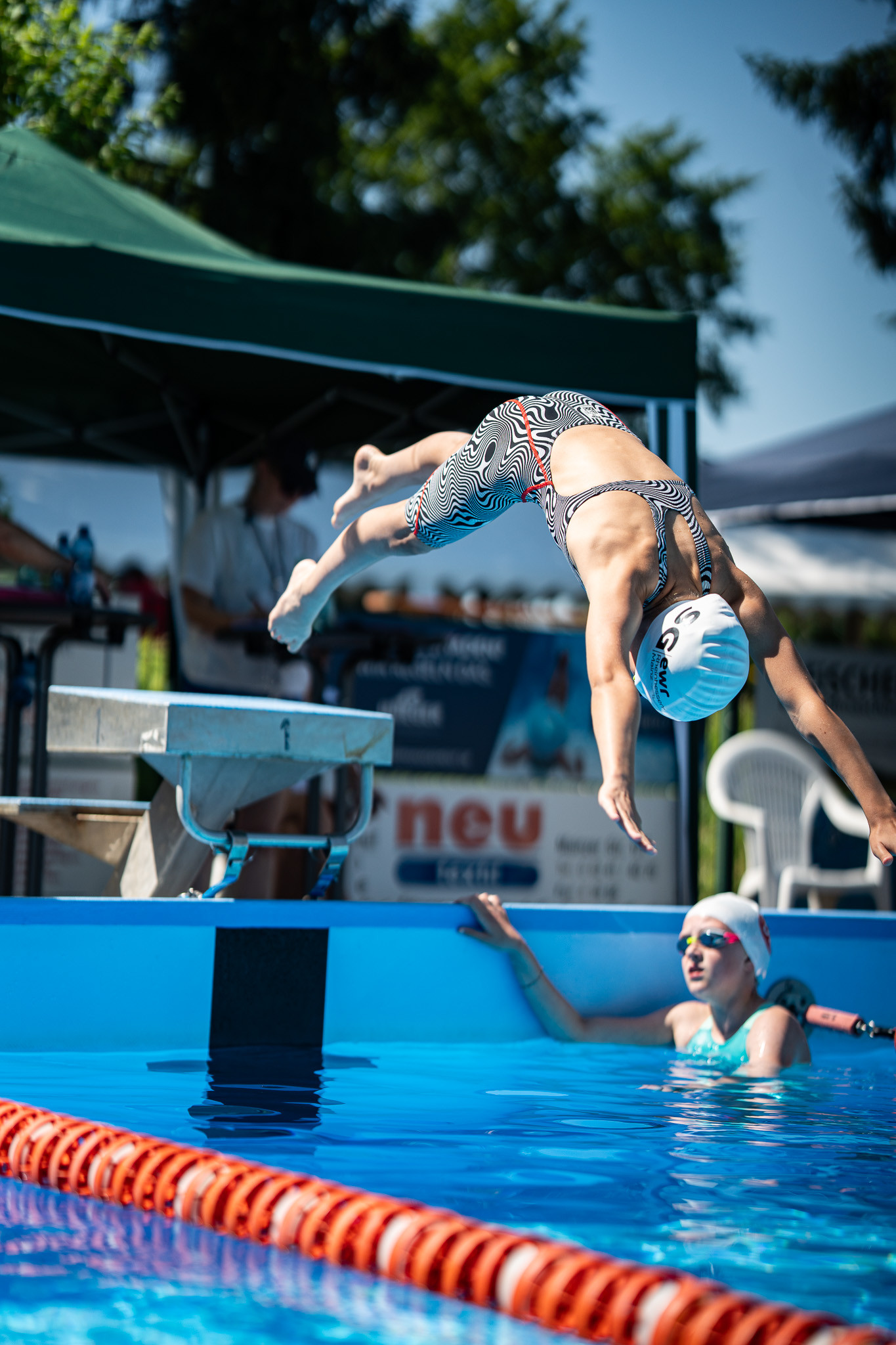Schwimmerin springt bei einem Schwimmwettkampf vom Startblock ins Wasser, während eine weitere Schwimmerin im Becken wartet.