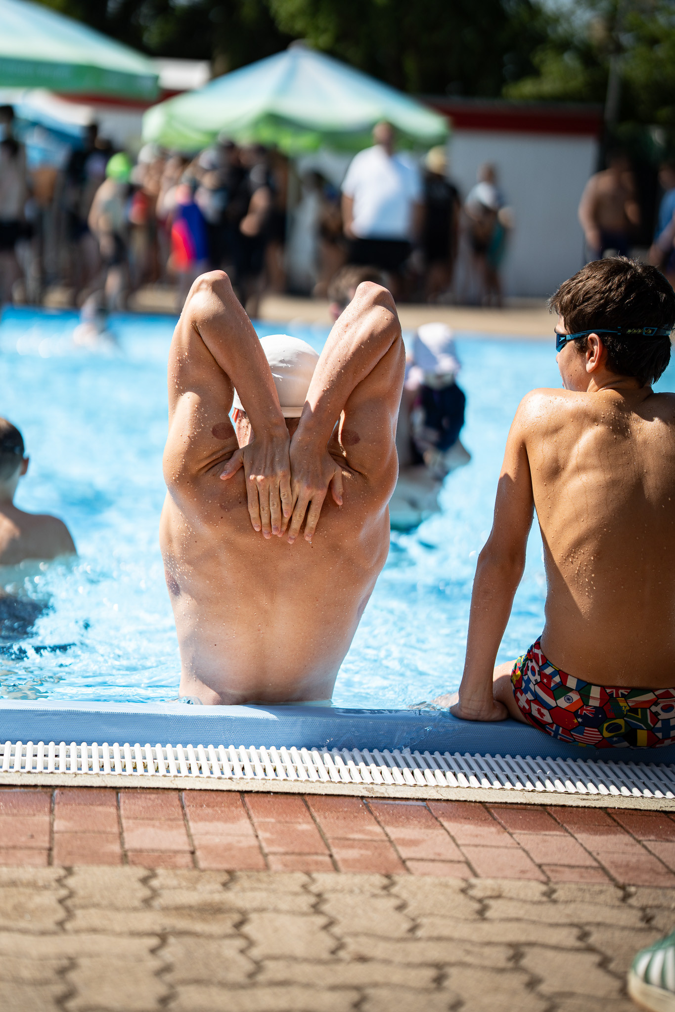 Zwei Jungen am Schwimmbecken, einer dehnt sich im Wasser, der andere sitzt am Beckenrand bei sonnigem Wetter