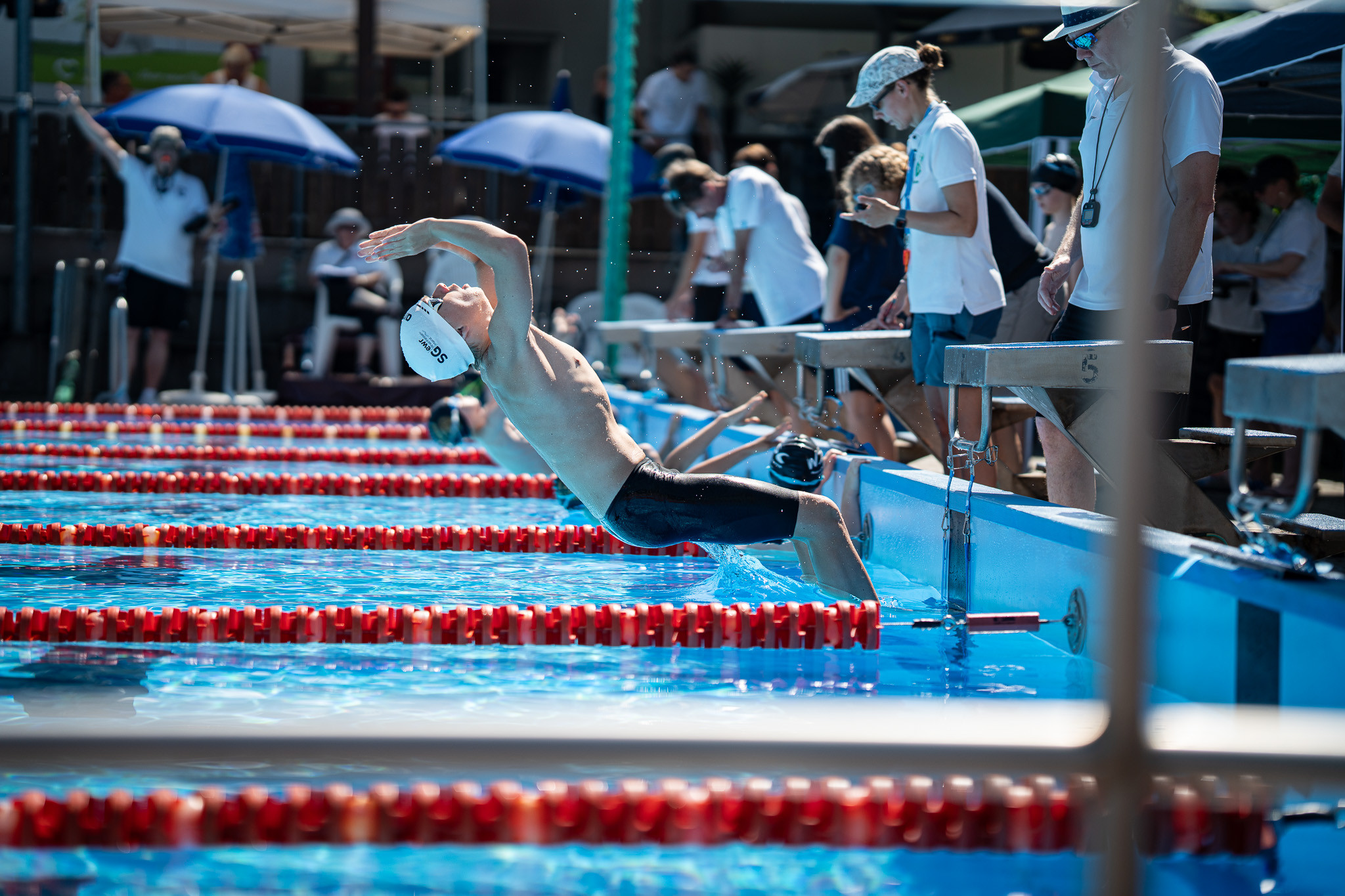Schwimmer beim Start eines Rückenschwimmrennens im Freibad mit Zuschauern und Kampfrichtern