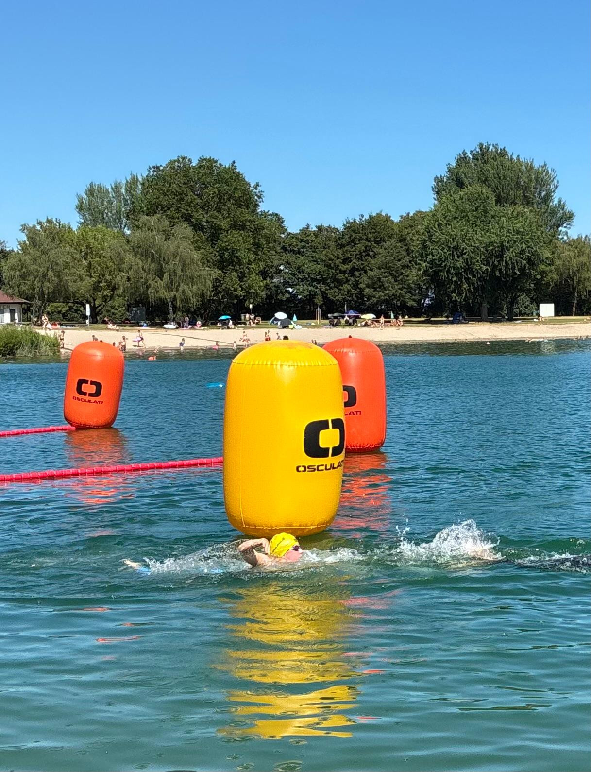 Schwimmer beim Freiwasserschwimmen mit gelber Badekappe und Bojen von Osculati an einem sonnigen Badesee