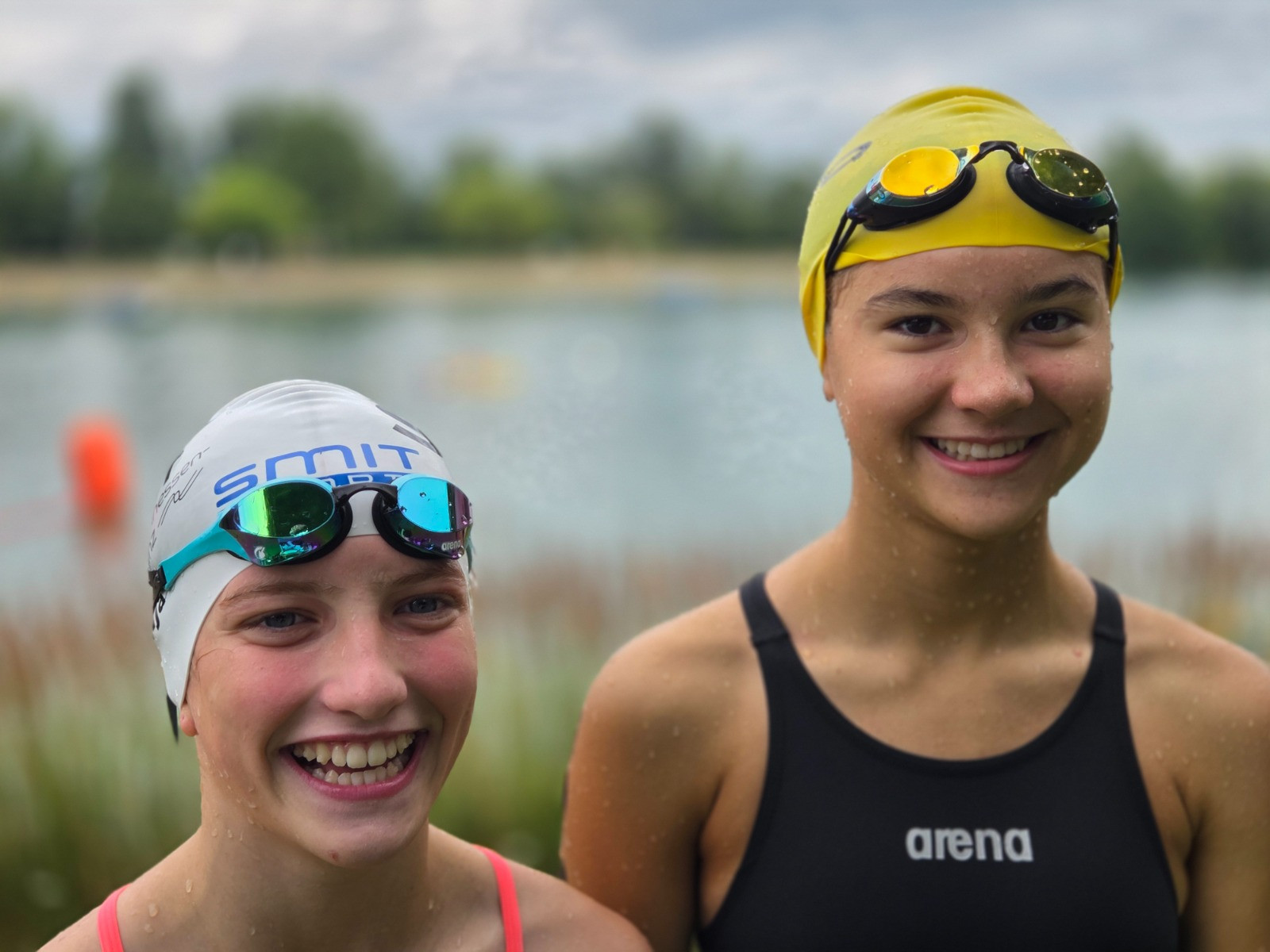 Zwei junge Schwimmerinnen mit Badekappen und Schwimmbrillen vor einem See beim Freiwasserschwimmen
