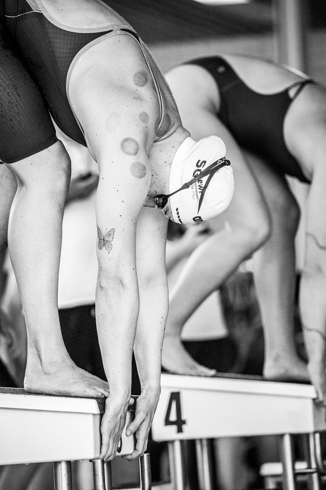 Schwimmerin am Startblock vor dem Wettkampf, schwarz-weiß, mit Schmetterling-Tattoo und Badekappe