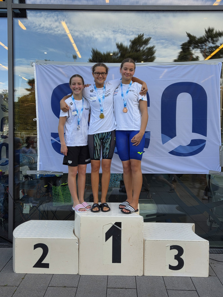 Siegerpodest bei Schwimmwettkampf mit Medaillengewinnerinnen und SGO-Banner Drei junge Schwimmerinnen mit Medaillen auf dem Siegerpodest bei einer Schwimmveranstaltung vor einem SGO-Banner.