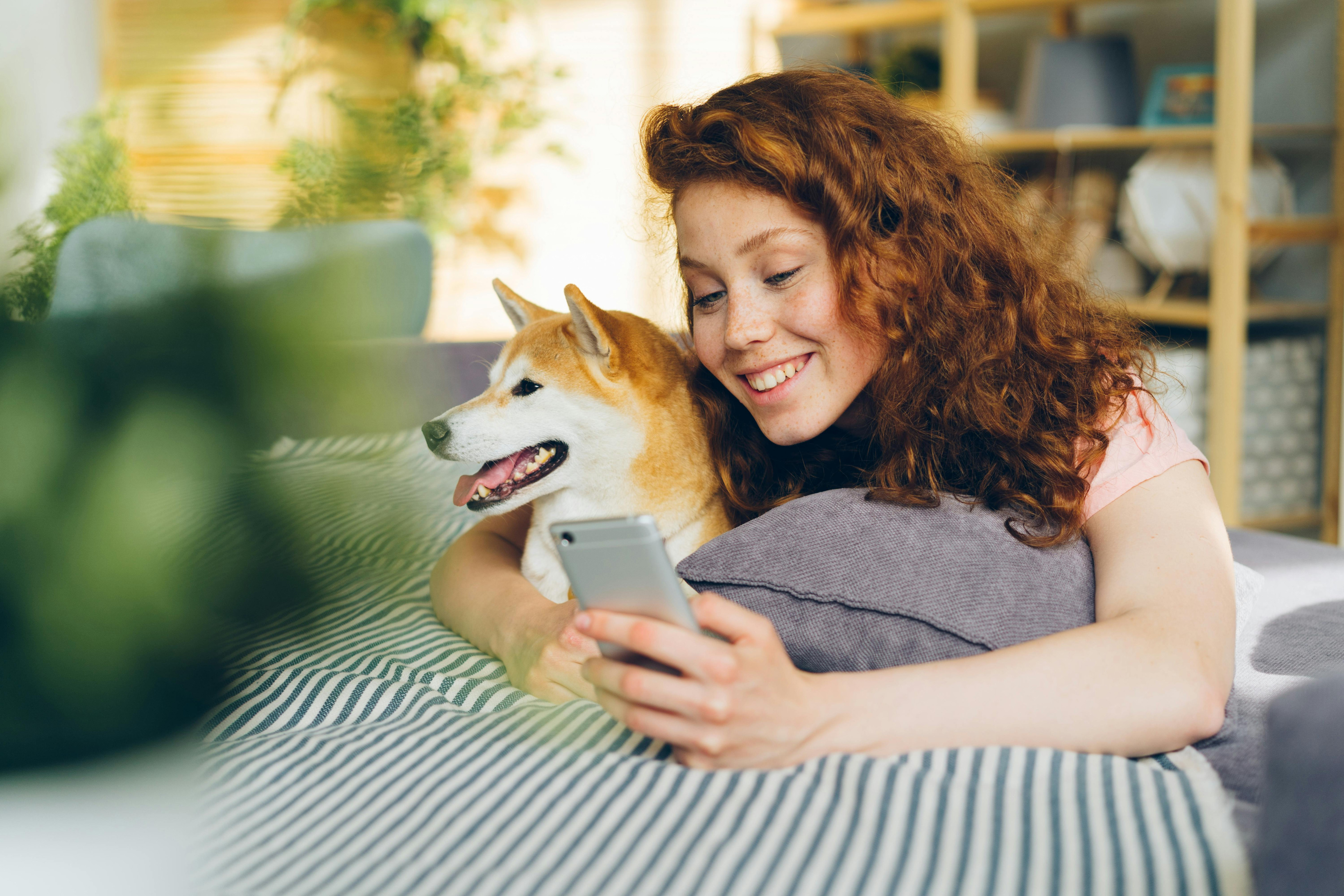 Frau mit Hund liegt auf dem Bett und nutzt ein Smartphone
