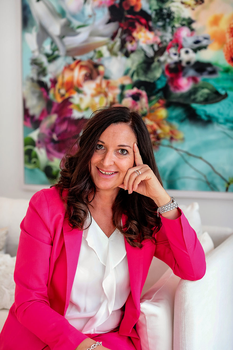 Katja Demming sitzt auf einem Sofa, trägt einen pinken Blazer und weißes Oberteil, im Hintergrund ein farbenfrohes Gemälde