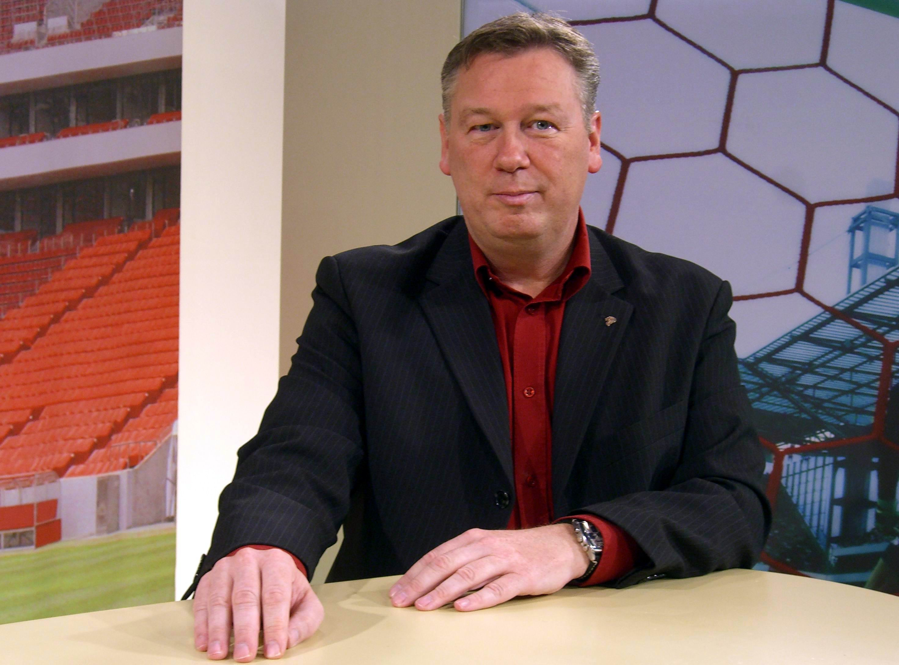 Ralf Friedrichs am Tisch im Studio, mit rotem Hemd und dunklem Sakko