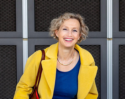 Schreiner Sabine mit gelbem Mantel und blauer Bluse vor moderner Fassade