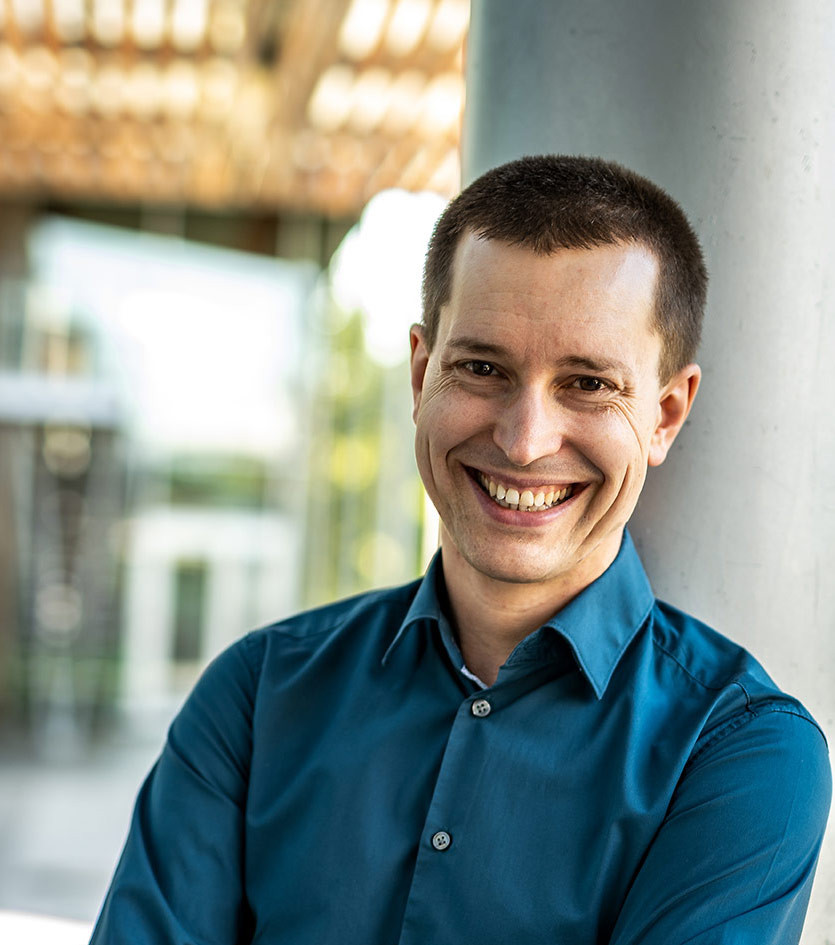 Portrait von Christian Koch, Mann im blauen Hemd, angelehnt an eine Säule