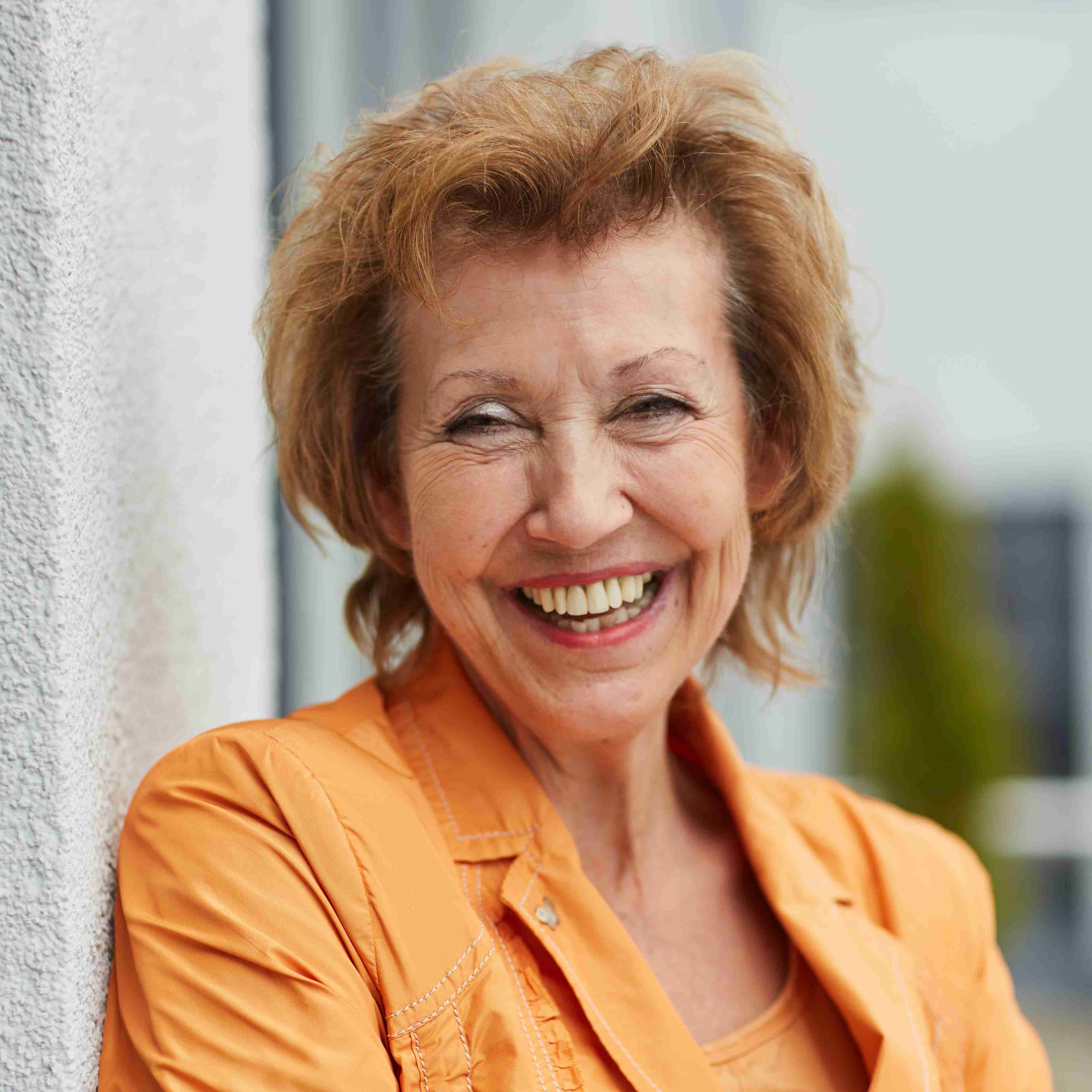 Porträt von Beate Löffler in orangefarbener Jacke, angelehnt an eine Wand