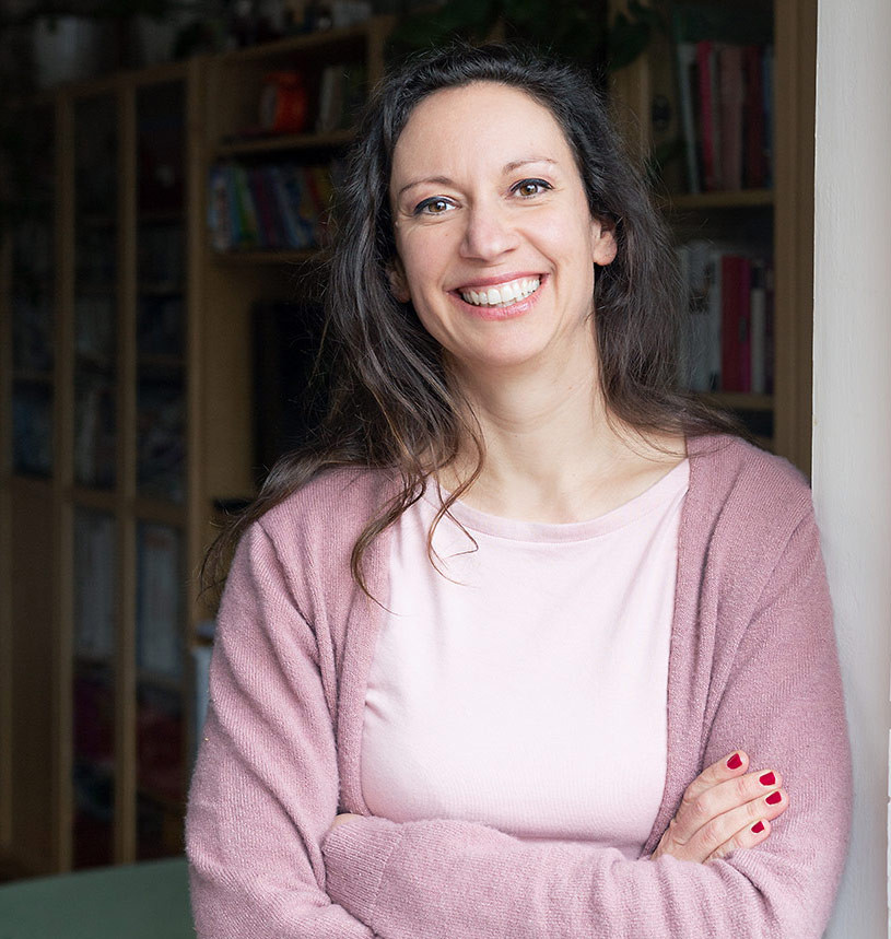 Portrait von Irene Rosinski mit verschränkten Armen in einem Raum mit Bücherregal im Hintergrund