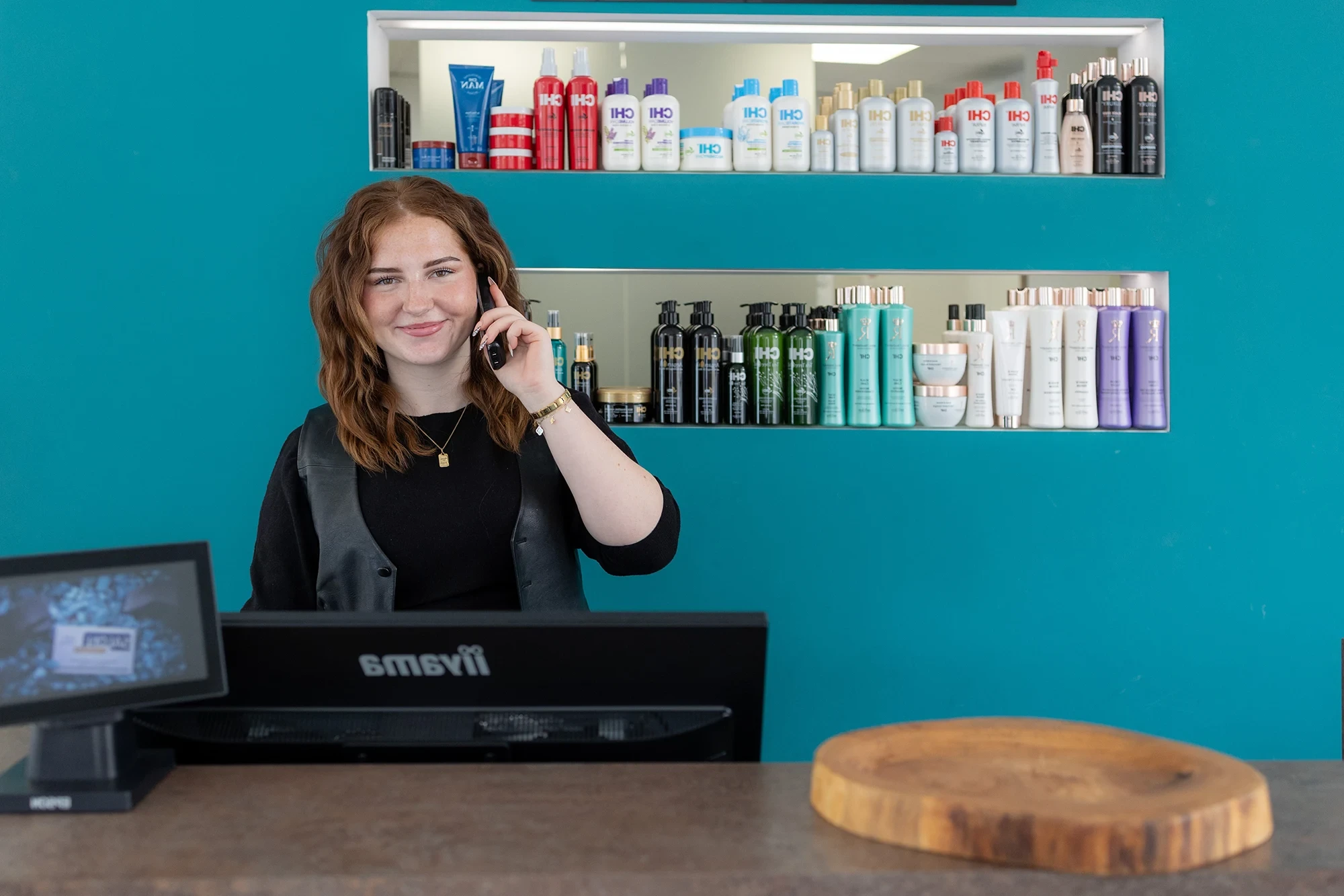 Headerbild Kontakt – Empfangsbereich eines Salons mit Mitarbeiterin am Telefon und Produkten im Hintergrund