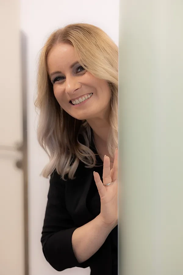 Jane lächelt und winkt freundlich hinter einer Wand hervor