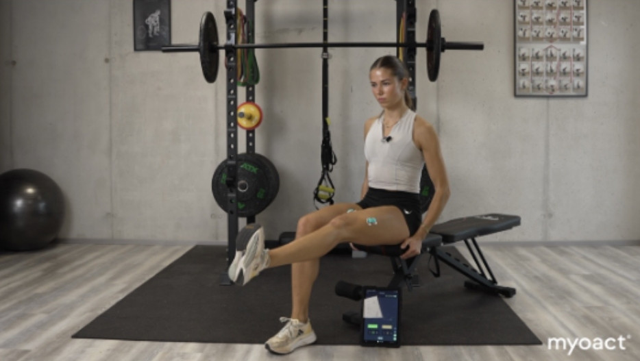 Frau macht Fitnessübung im Fitnessstudio mit Asana-App auf Tablet, Hantelbank und Gewichte im Hintergrund