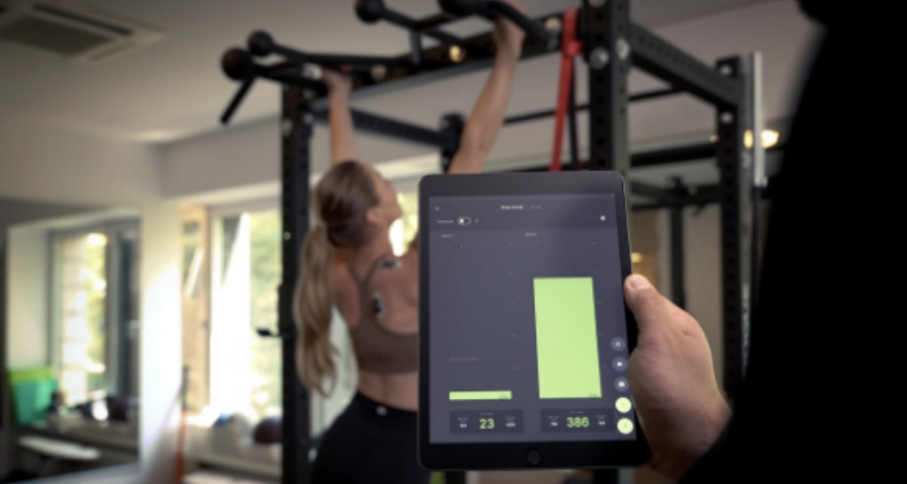 Bildschirmfoto der App Asana.com auf einem Tablet, im Vordergrund, während im Hintergrund eine Frau Klimmzüge macht
