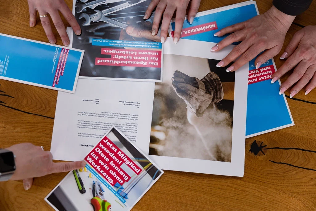Mehrere Hände betrachten verschiedene Werbemittel und Flyer auf einem Tisch, die sich auf Handwerk und Mitgliedschaft beziehen.