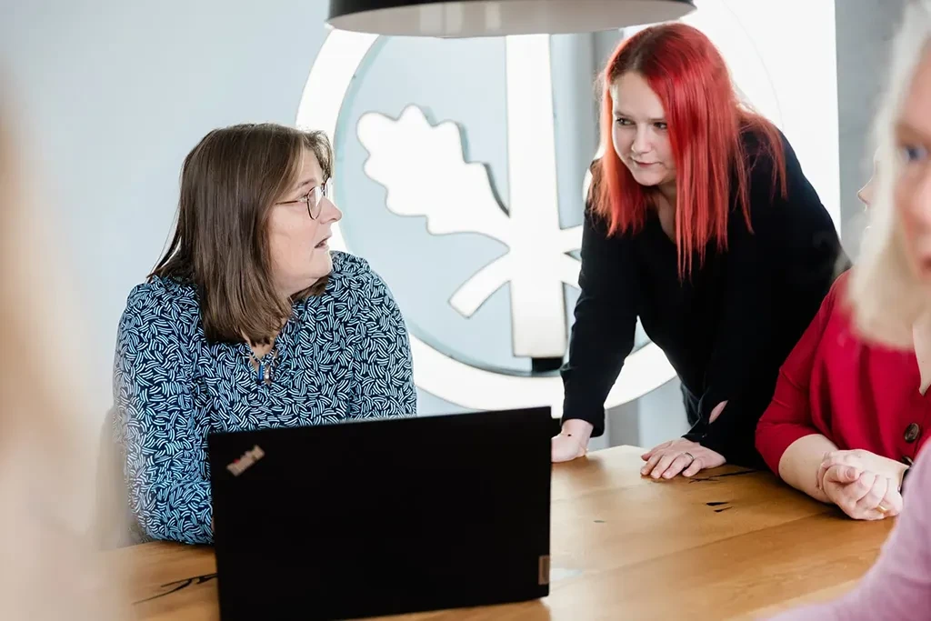 Zwei Kolleginnen besprechen etwas am Schreibtisch im Büro, eine sitzt am Laptop, die andere steht daneben.